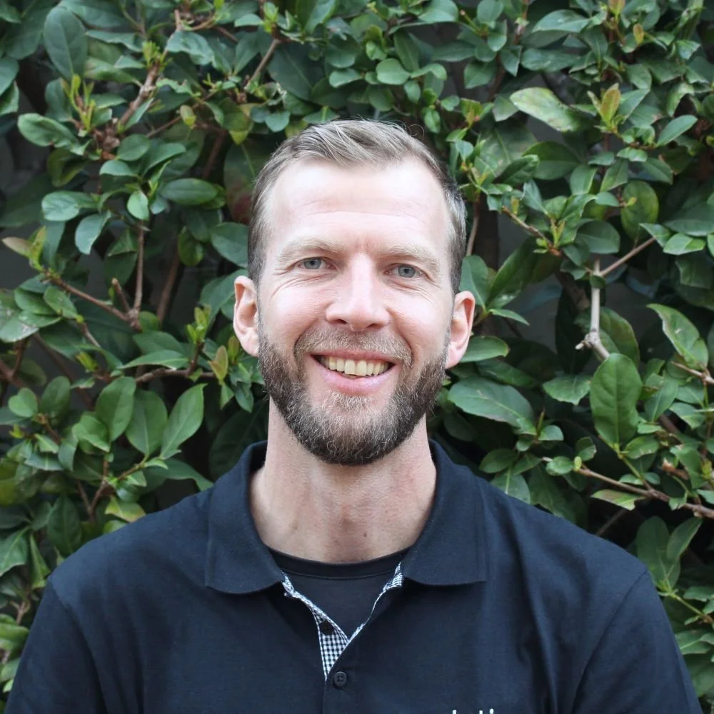 A man with short light brown hair and a beard smiling in front of green foliage.