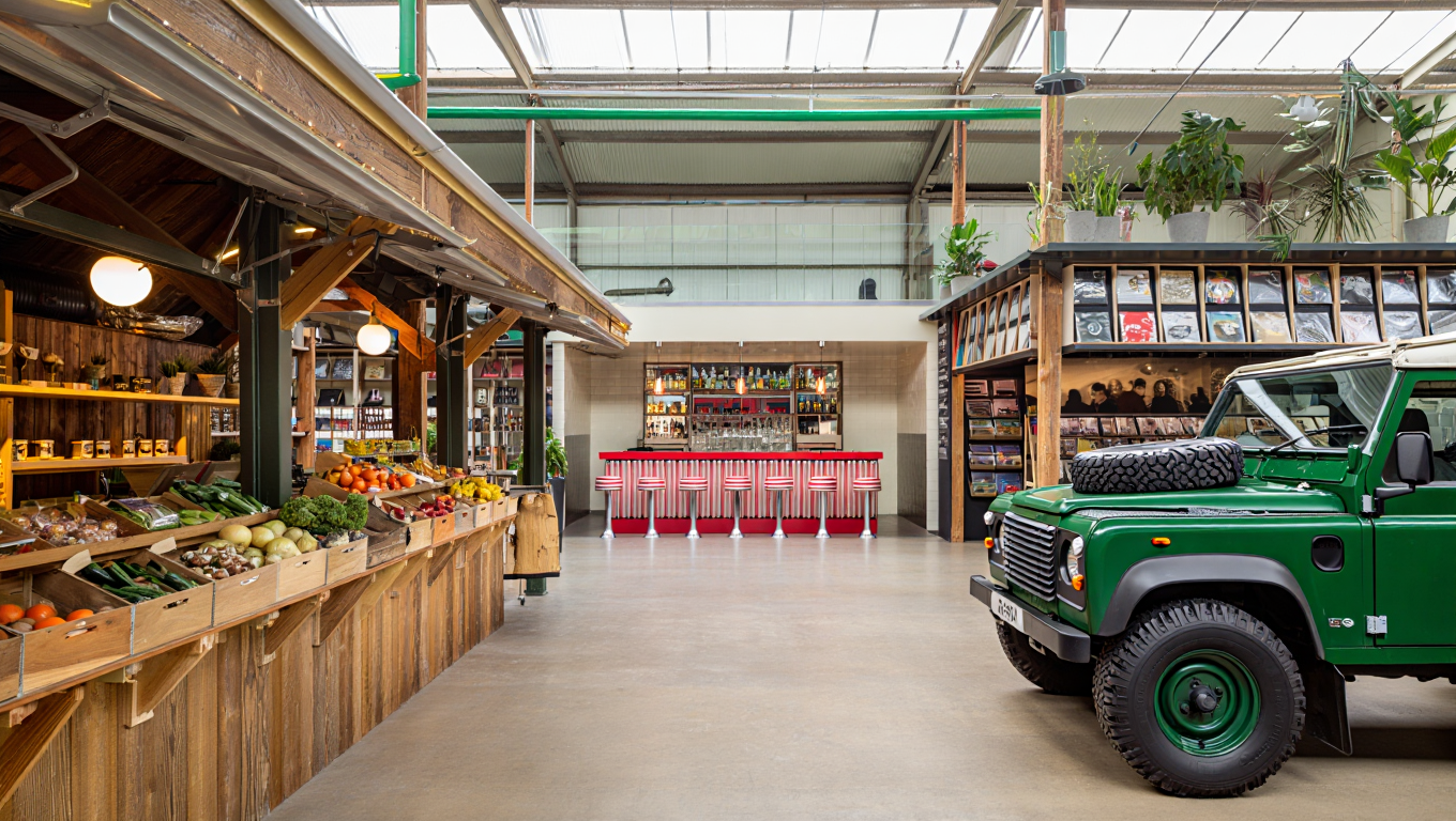 Interior of a farm shop with fresh produce on wooden shelves, a vintage green off-road vehicle, and a bar area with red-and-white bar stools.
