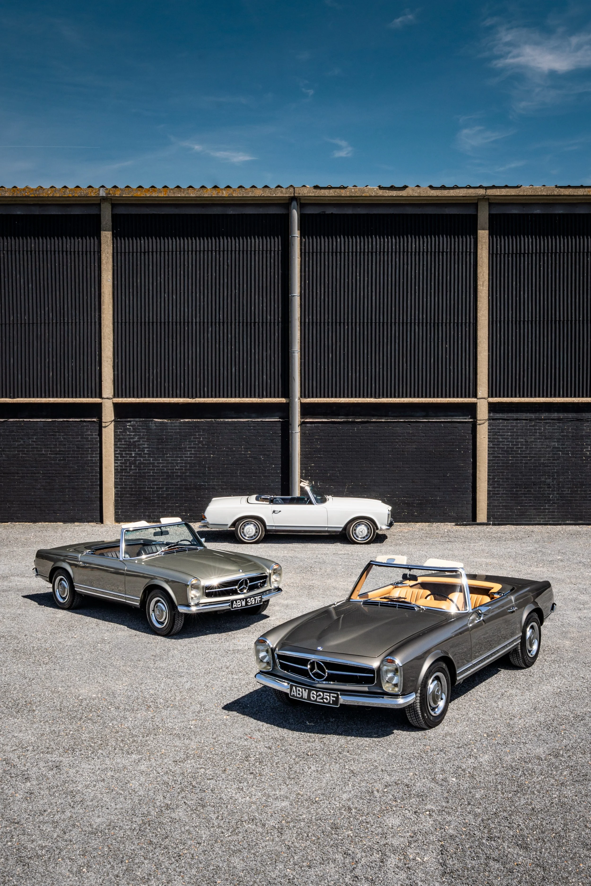 Four vintage Mercedes-Benz convertibles parked in a gravel lot in front of a black industrial building with a corrugated metal upper section and concrete lower section, under a partly cloudy blue sky.