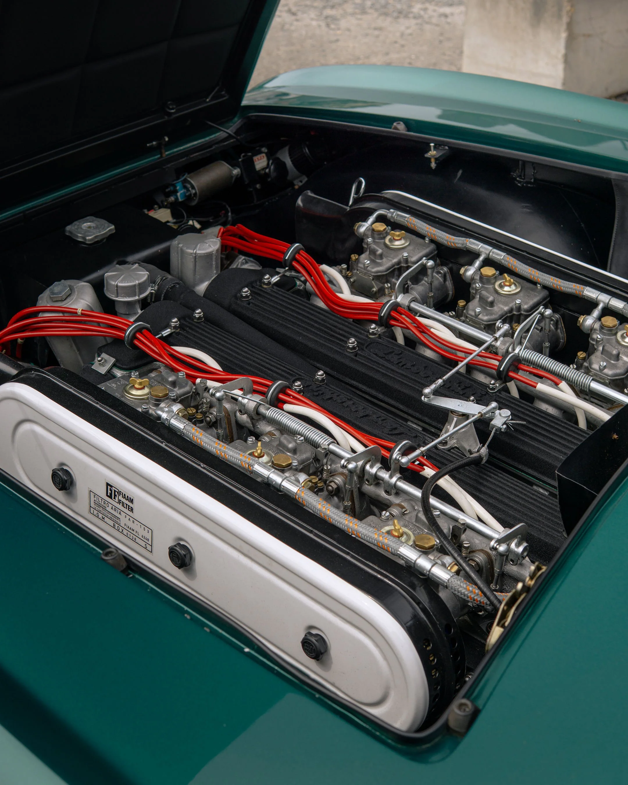 Close-up of a vintage car engine bay with black, silver, and red components, visible hoses, and mechanical parts.