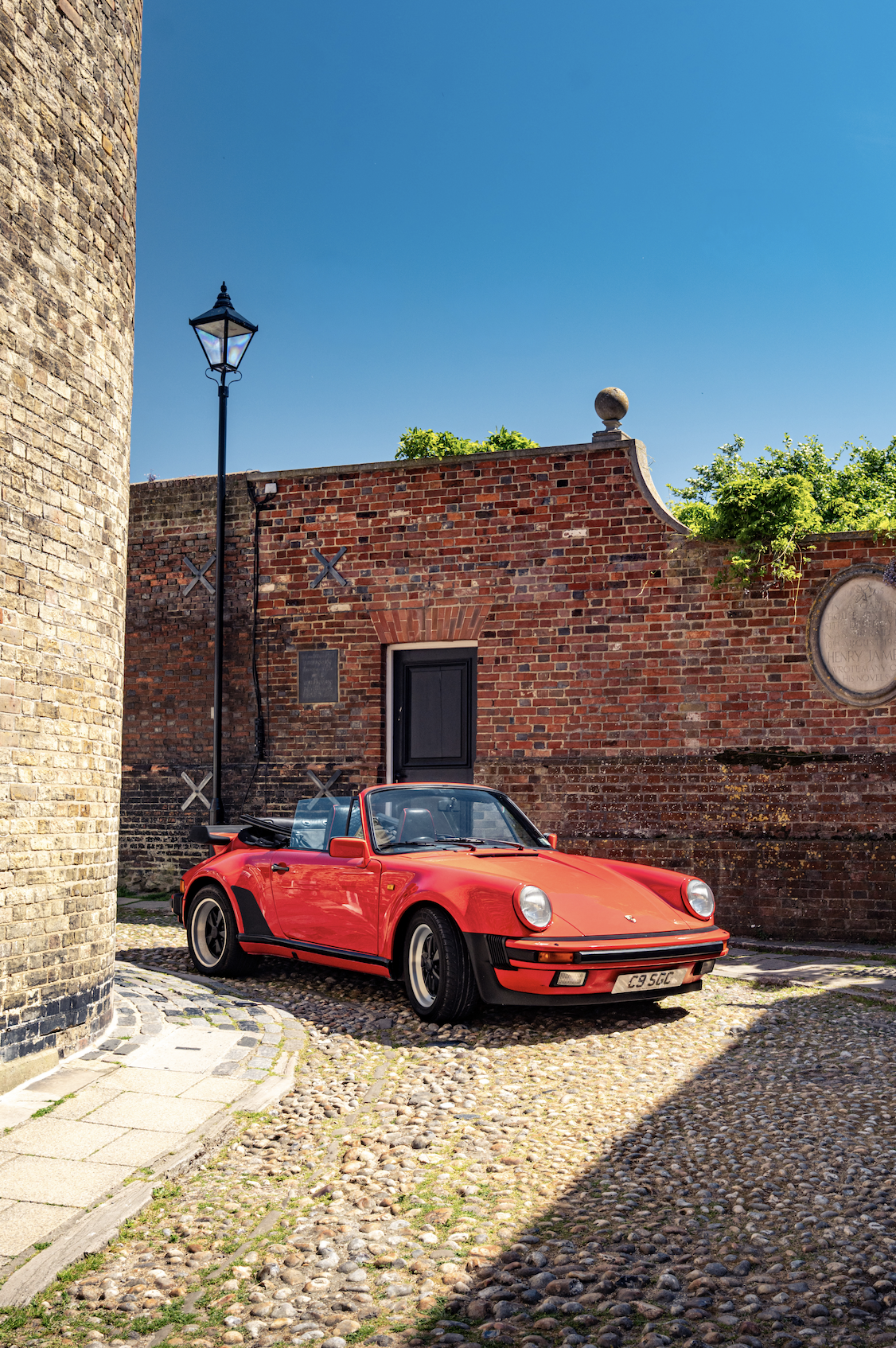 A red Porsche convertible parked on a cobblestone street in front of a brick wall, with a lamp post and a stone corner on the left. The sky is clear and blue.