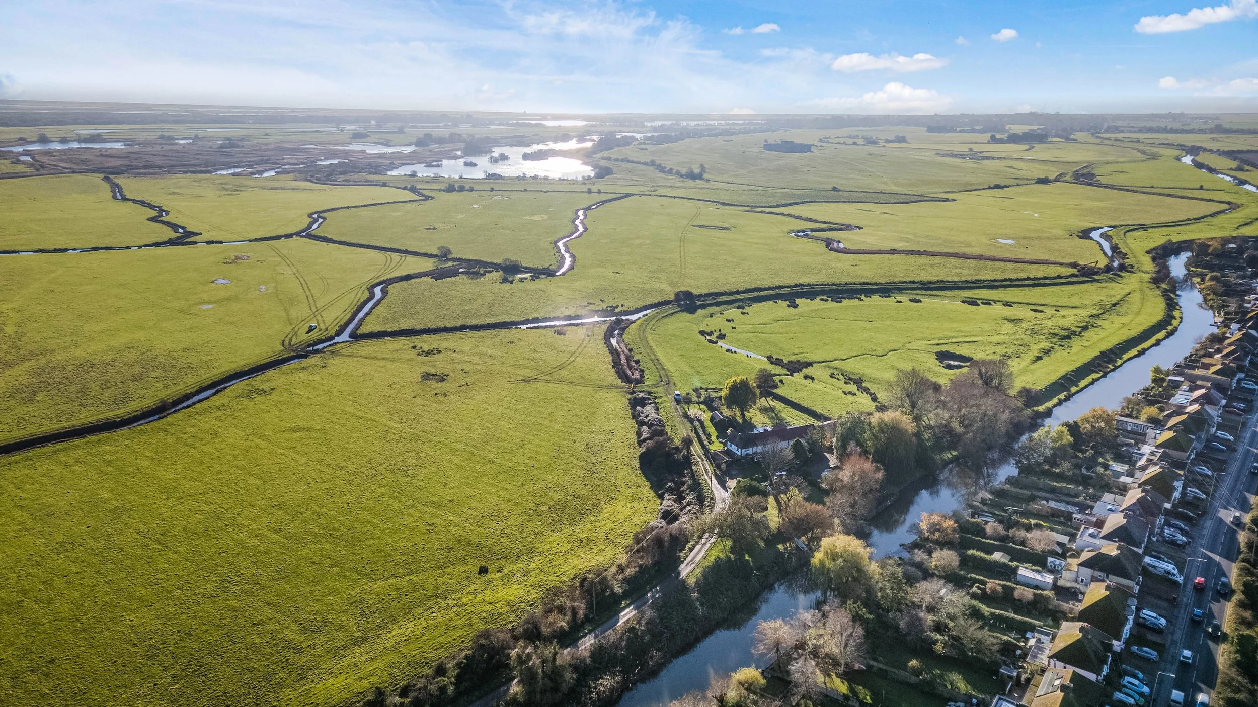 Aerial view of a green rural landscape with fields, meandering waterways, and a row of houses along a street.