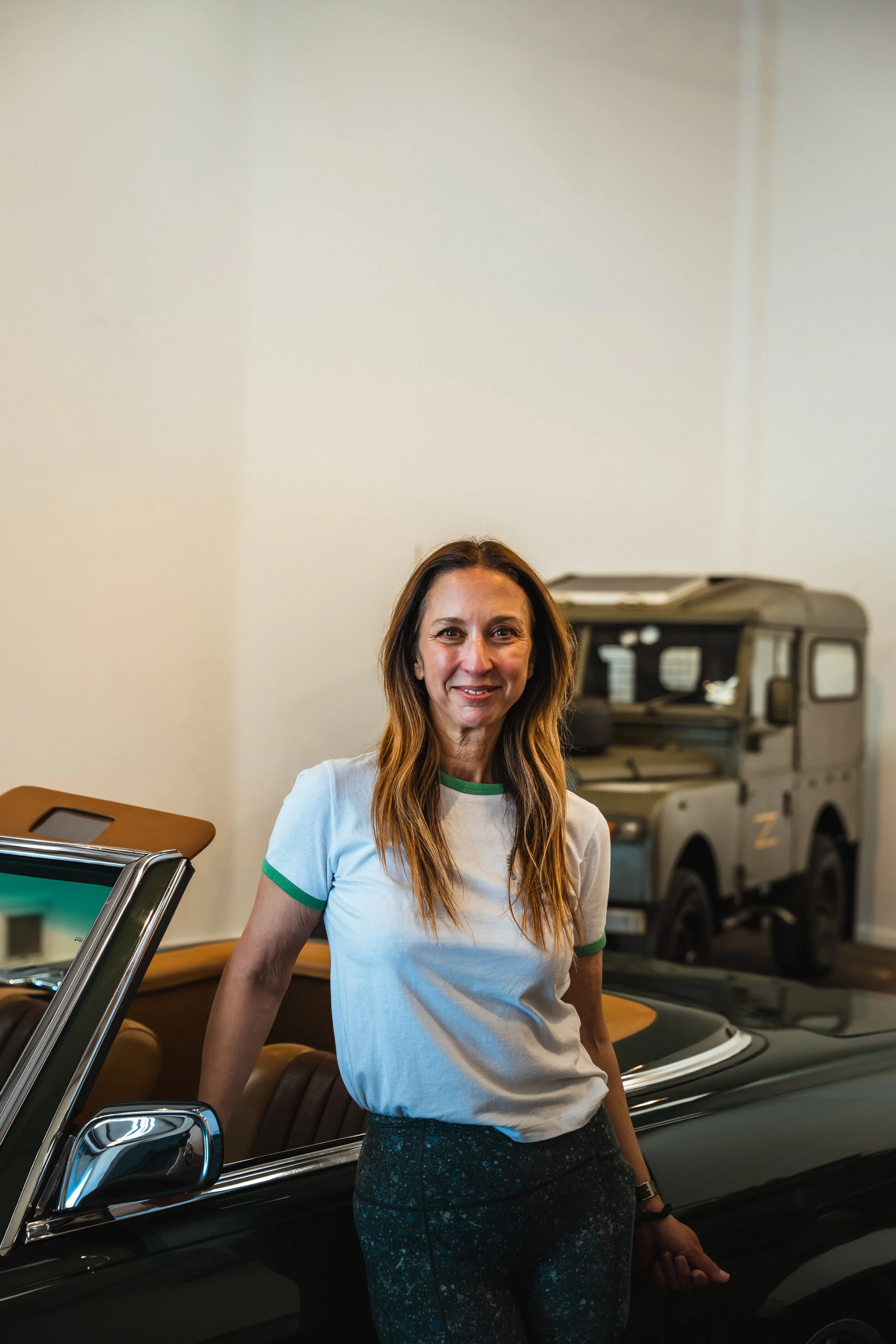 Woman with long hair in a white and green t-shirt standing next to a black classic car.