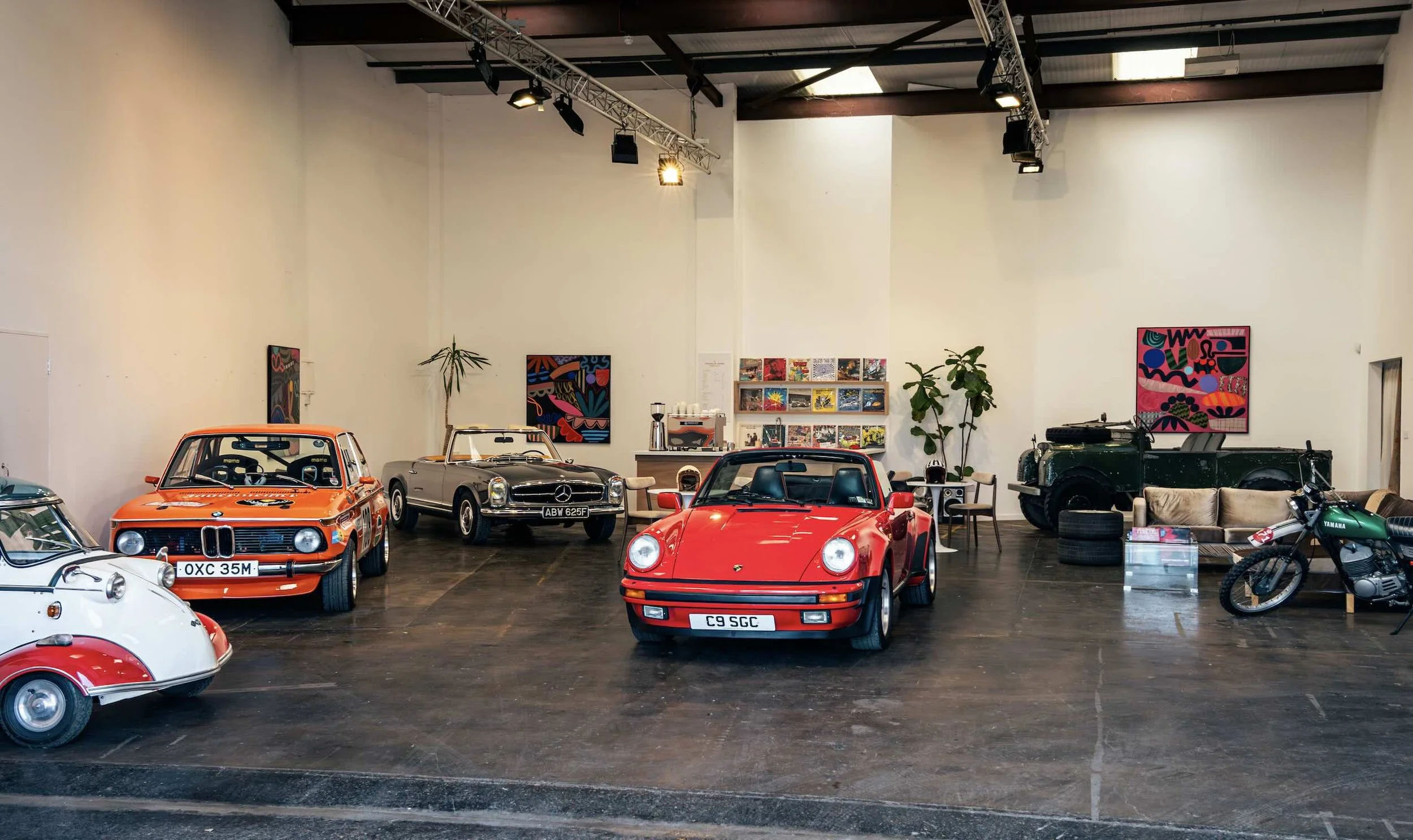 Inside a showroom with vintage and sports cars, including a red Porsche 911, an orange racing car, a Mercedes-Benz, and motorcycles, with colorful artwork on the walls and indoor plants.