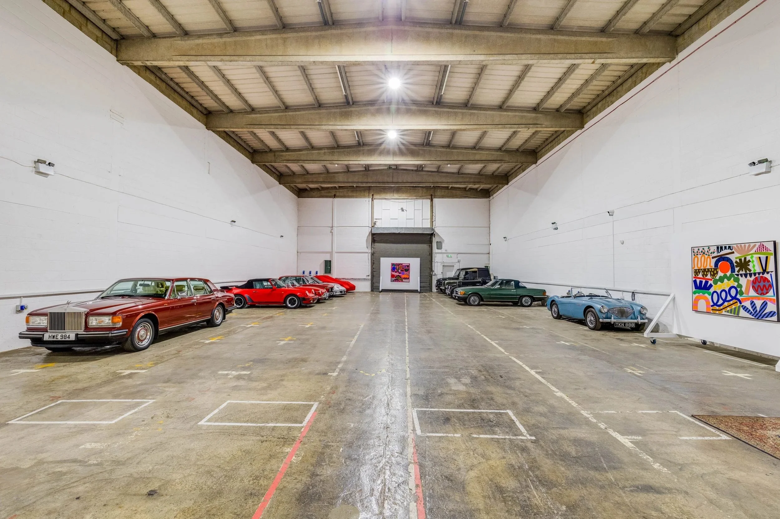 An indoor industrial space with classic and vintage cars parked along the sides, artwork displayed on the right wall, and a large roller door at the back.