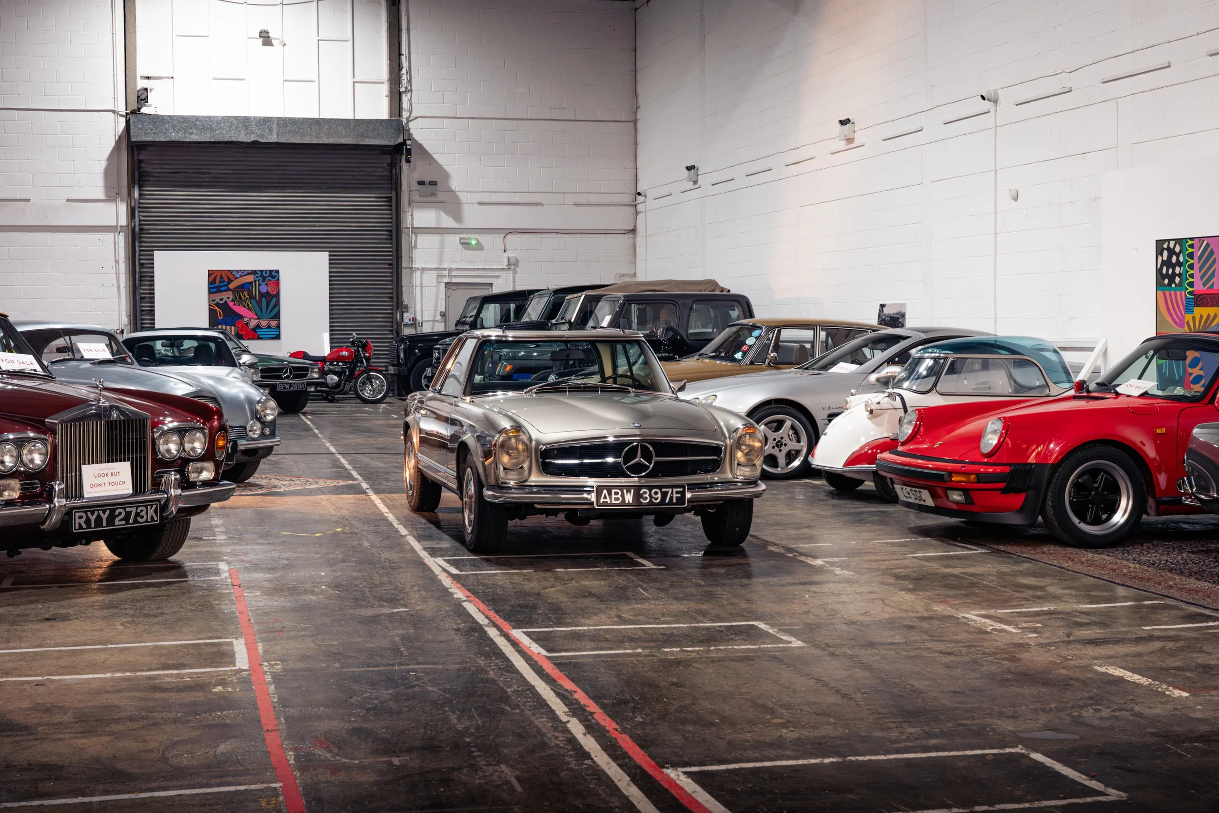 Indoor warehouse with a collection of vintage and classic cars, including a red sports car, silver Mercedes-Benz, and other historic vehicles, with artwork on the walls.
