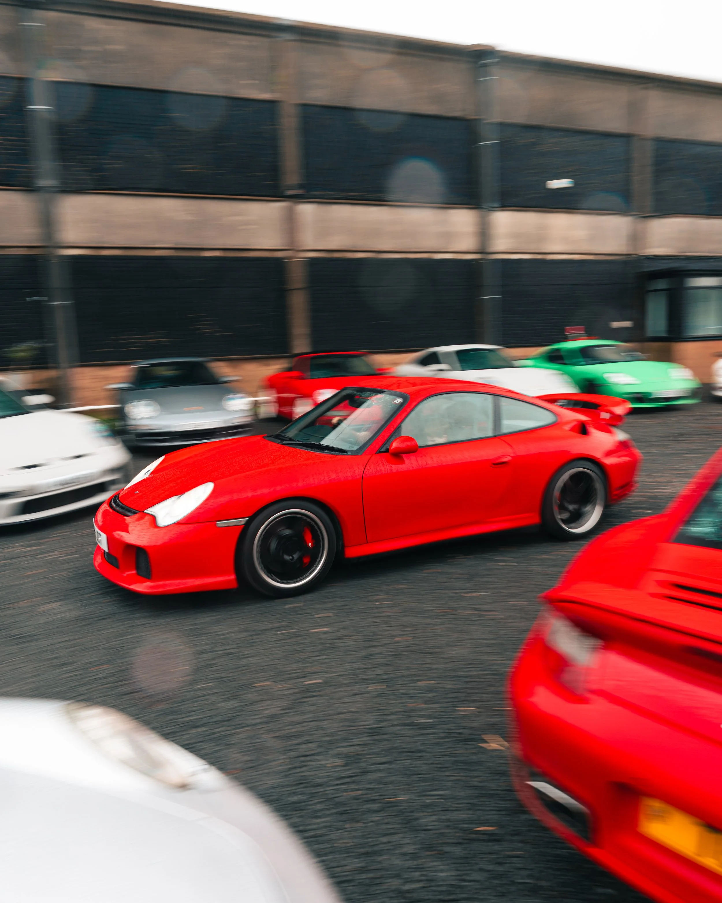 Blurred image of several parked sports cars, including a red car in the foreground, outside a building with black and brown facade.