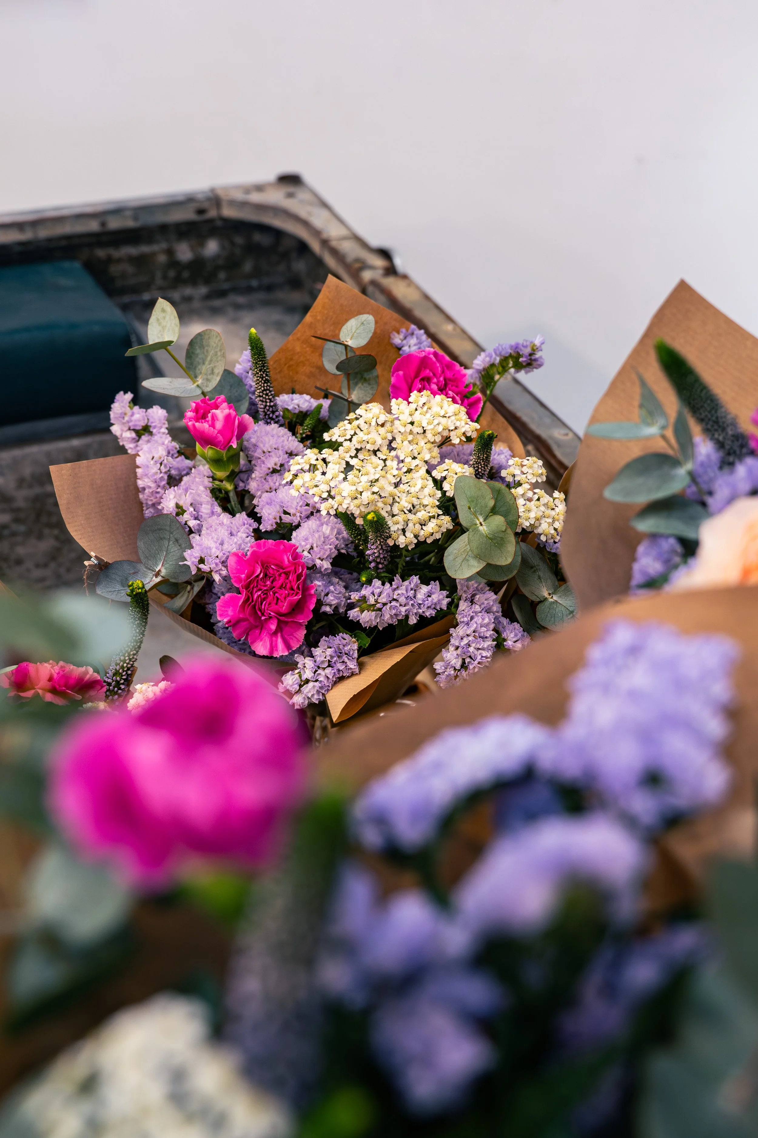 A bouquet of colorful flowers, including pink, white, and purple blossoms with eucalyptus leaves, wrapped in brown paper, placed on a surface near a metal container.