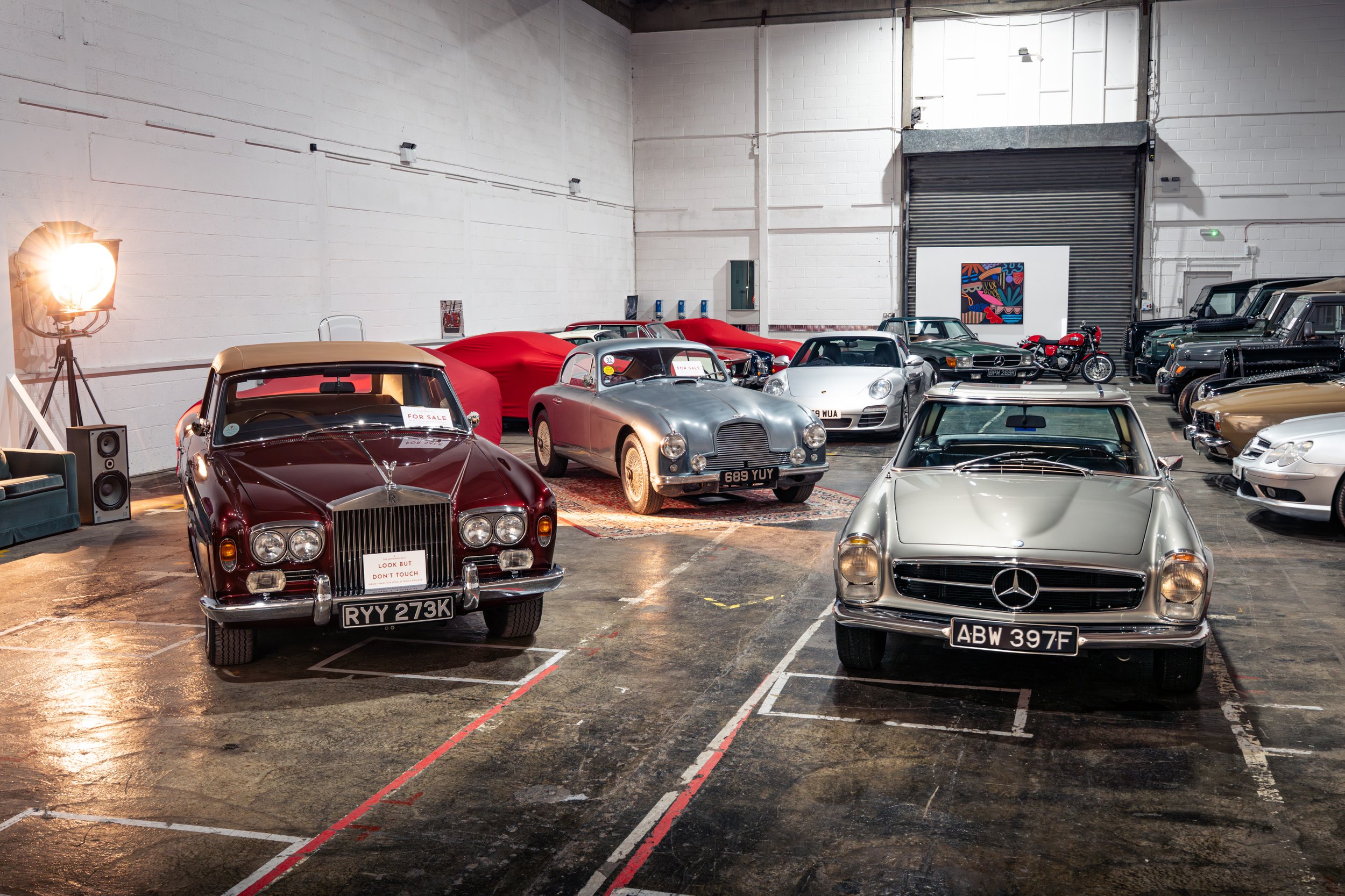 Multiple classic and luxury cars parked inside a spacious garage or showroom, with some covered by red cloths and a motorcycle among them, featuring white walls, a roller door at the back, artwork, and vintage-style lighting.