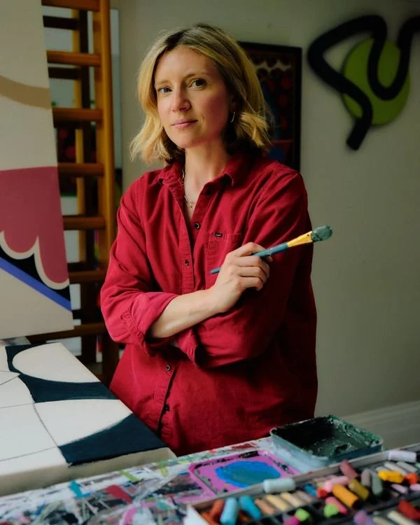 A woman with blonde hair, wearing a red shirt, holding a paintbrush, standing in front of colorful artwork and art supplies on a table.