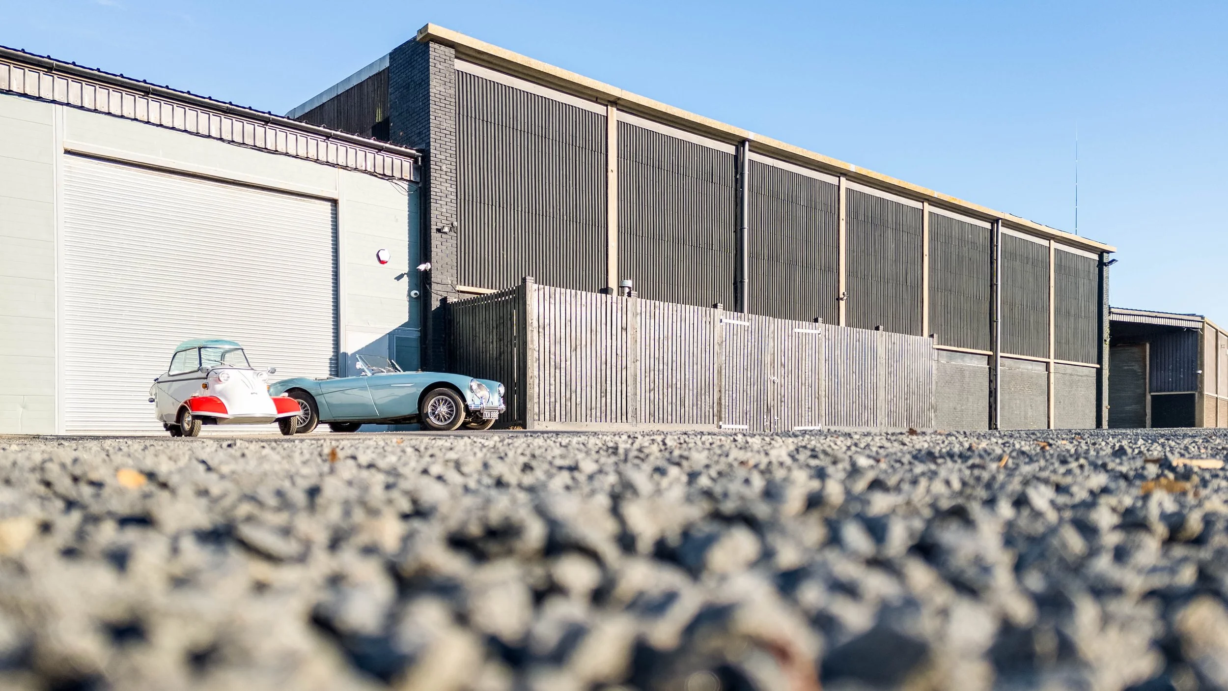 Two vintage cars, one silver and one white with red accents, parked in front of an industrial building with large gray and black walls and a closed garage door.