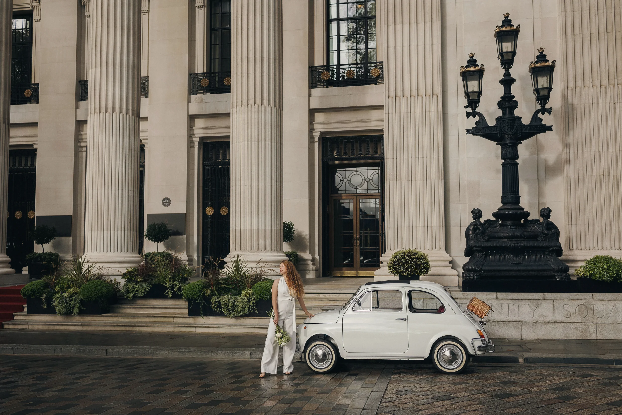A woman in a white outfit standing beside a white vintage Fiat 500 parked in front of a grand building with tall columns and ornate lamps, holding a bouquet of flowers.