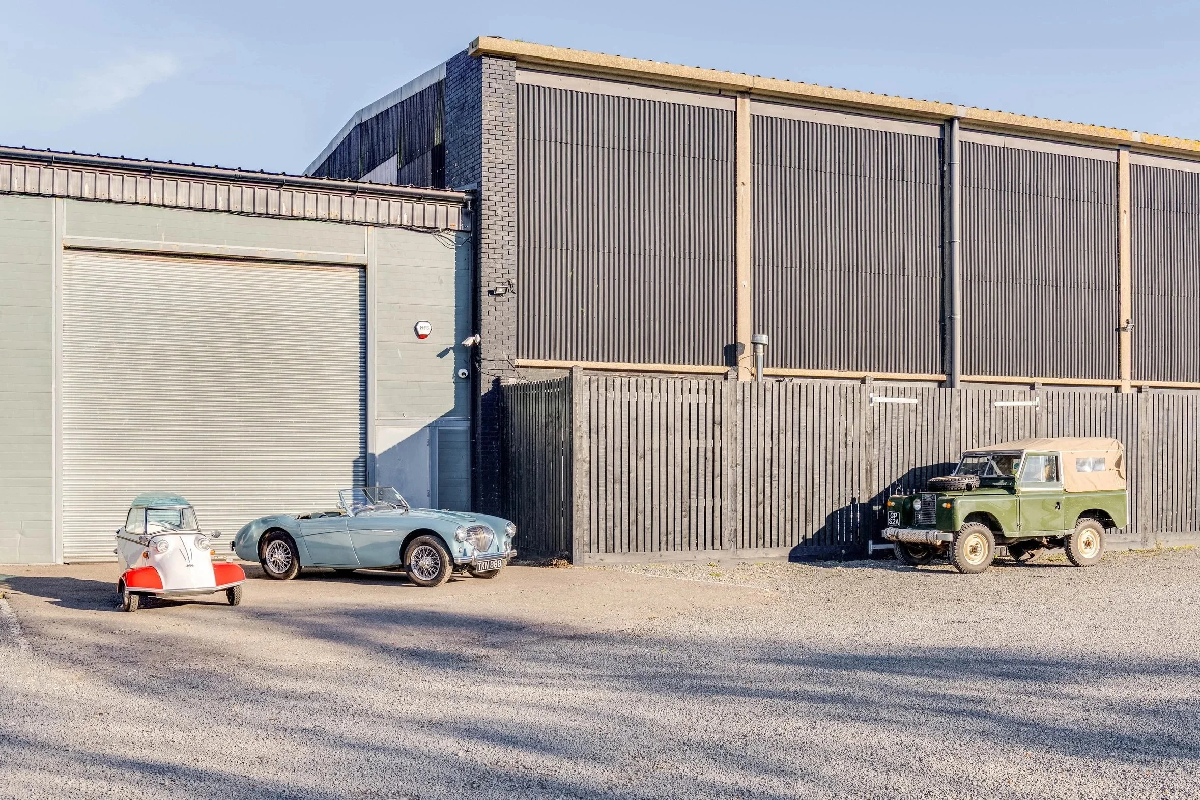 Three vintage cars parked in front of industrial buildings, one small white tow vehicle, one light blue convertible, and one green off-road vehicle, under a clear sky.