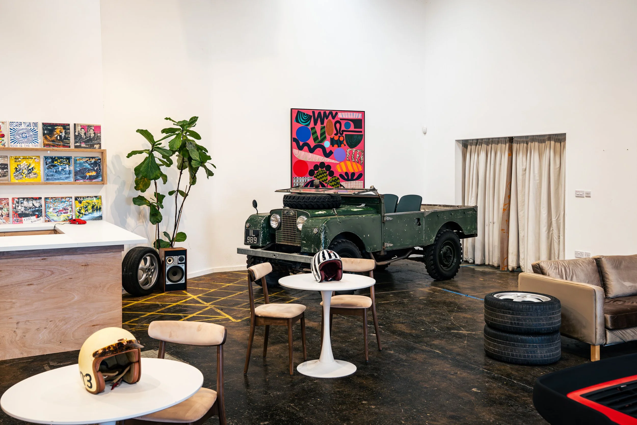 An indoor space with a vintage green Land Rover vehicle, a wall painting of colorful abstract shapes, a bookshelf with vinyl records, a potted plant, tables and chairs, racing helmets on the table, and stacked tires.