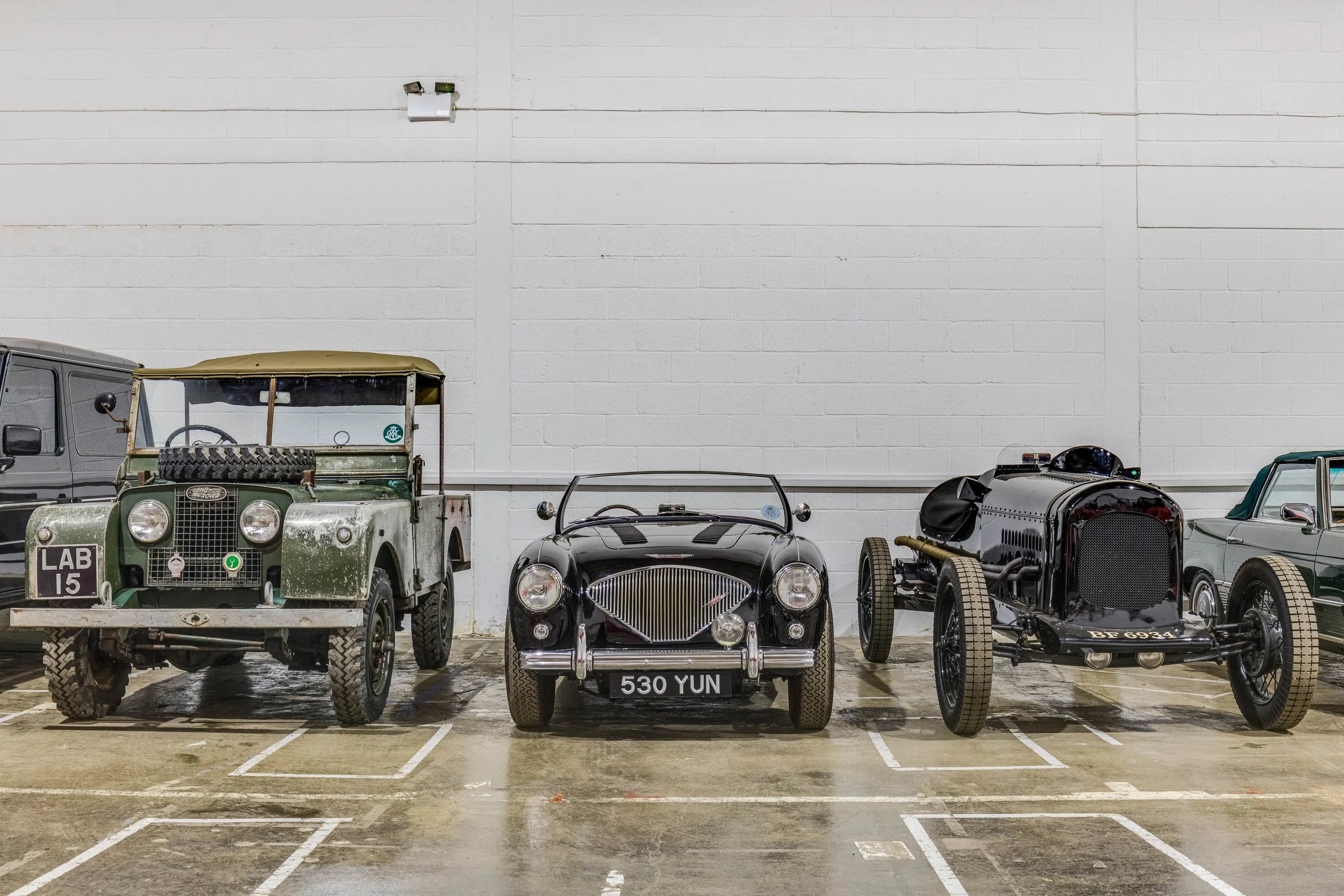 Three vintage cars parked in a row inside a garage with a white brick wall.