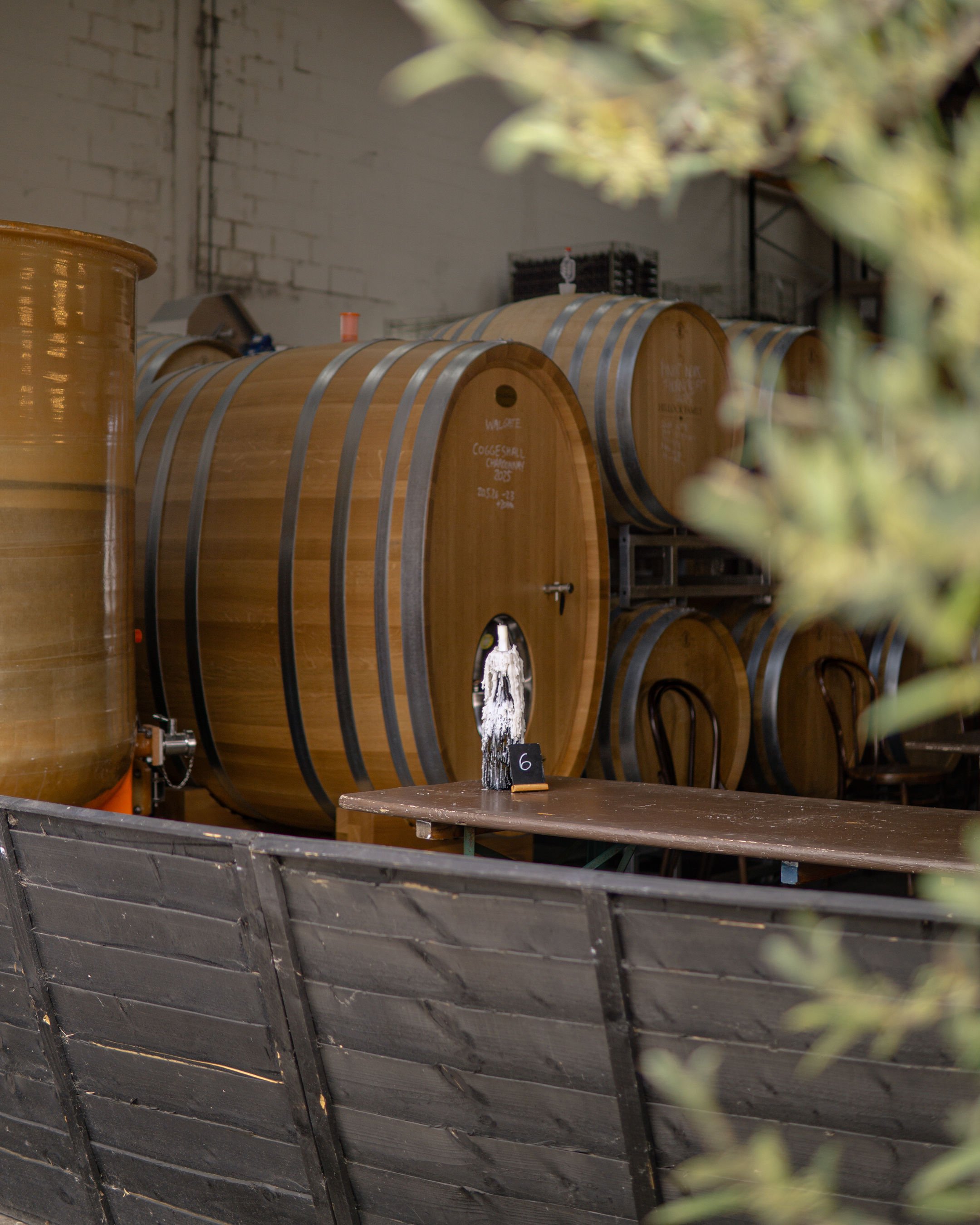 Wooden wine barrels stored in a warehouse or cellar with a brick wall in the background, some items on top of the barrels, and a black metal rack.