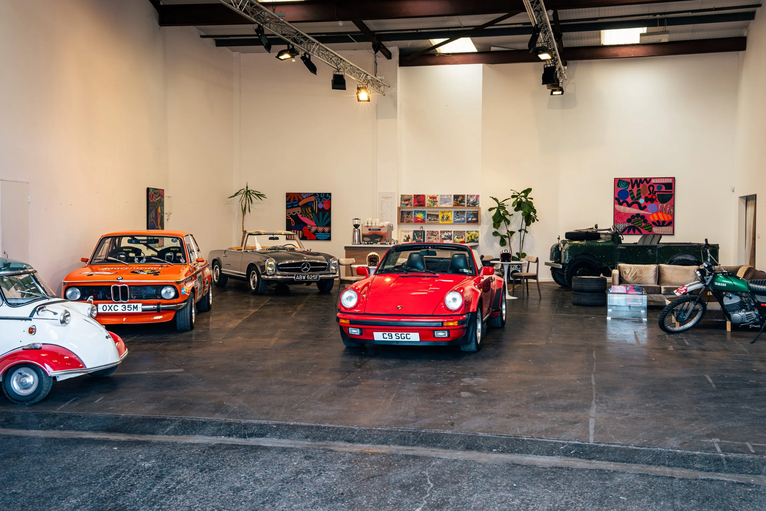 An indoor coworking space and market hall with several classic cars displayed, including a red Porsche, an orange BMW race car, and a silver Mercedes-Benz convertible, with artworks on the walls and some furniture and plants in the background.