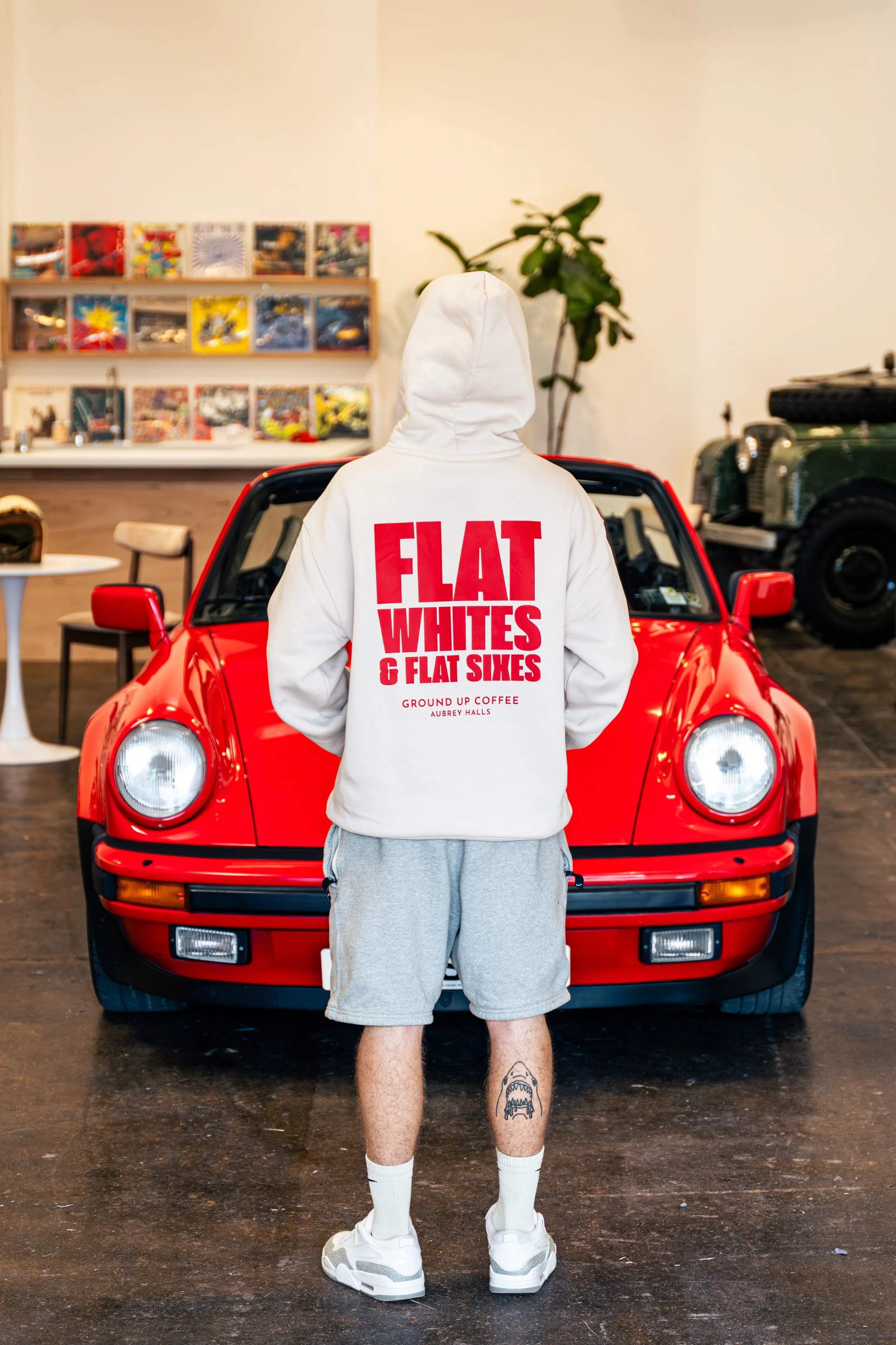 Person standing in front of a red sports car, facing away, wearing a white hoodie with bold red text on the back, gray shorts, white sneakers, and white socks. Inner walls decorated with magazines or comics, a wooden shelf, a vintage black car, plants, and a small table with a helmet.