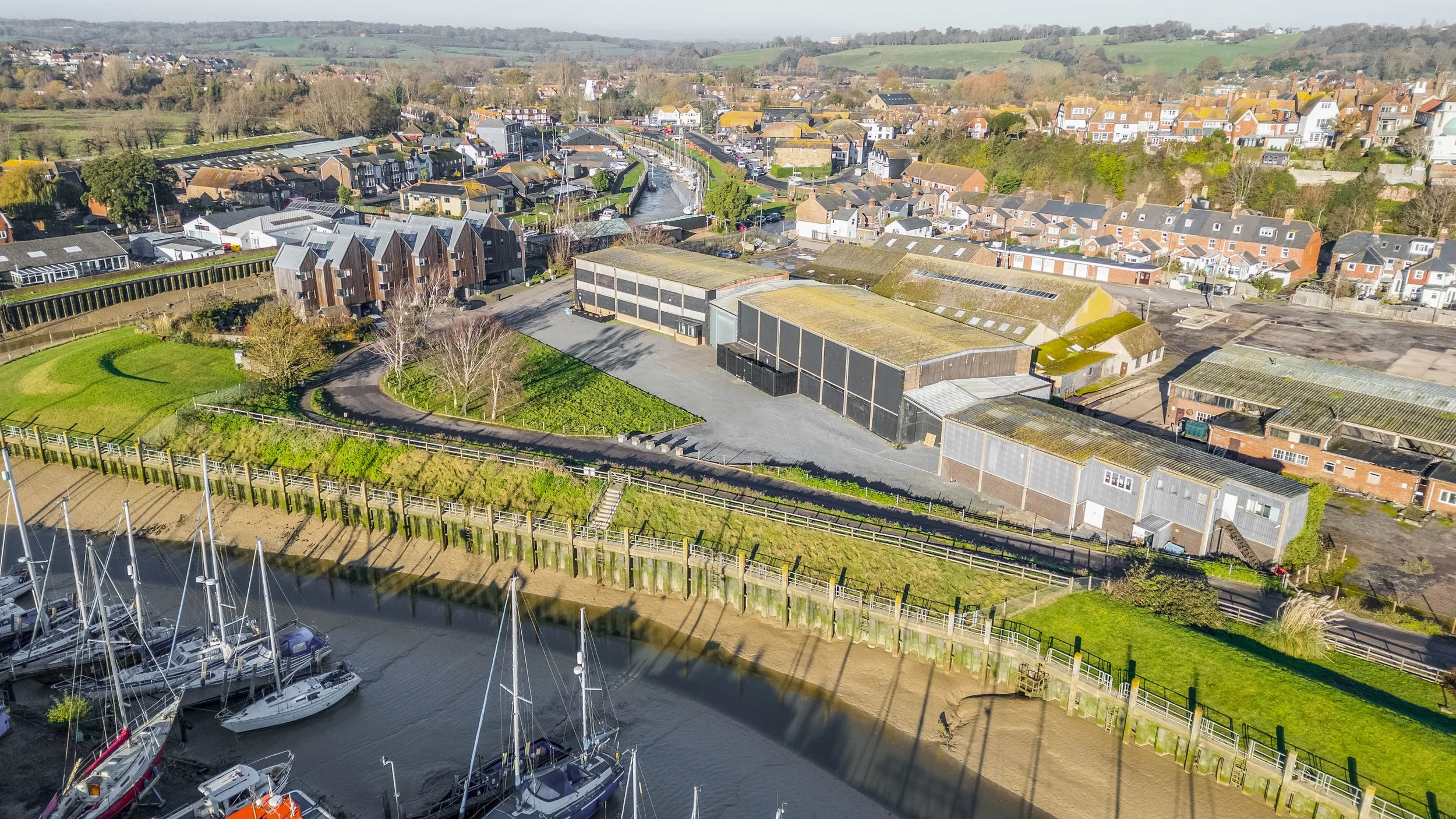 Aerial view of a harbor with sailboats, a town with residential houses, commercial buildings, green spaces, and rolling hills in the background.
