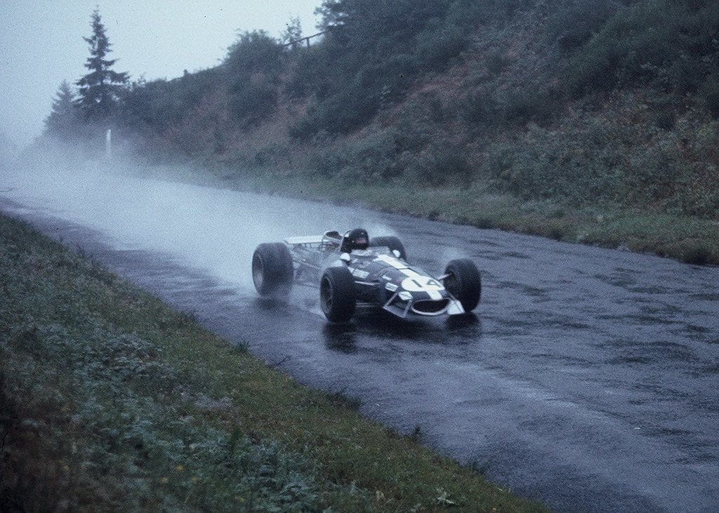 A vintage race car speeding on a wet, cloudy road in a rural area with trees and hills.