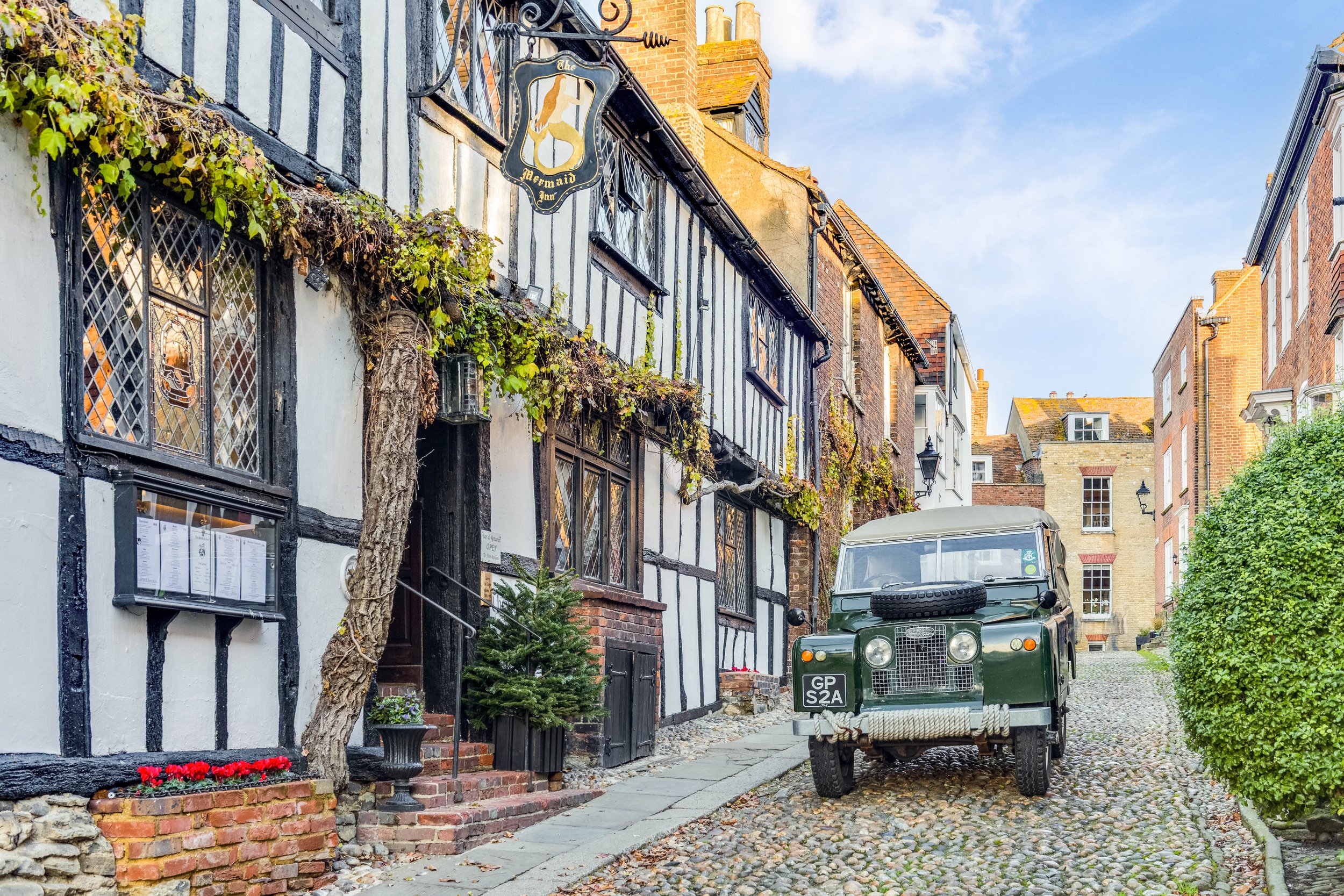 A narrow cobblestone street with a vintage green utility vehicle parked on the side, in front of buildings with black and white timber framing and brick facades, and greenery hanging from the buildings.