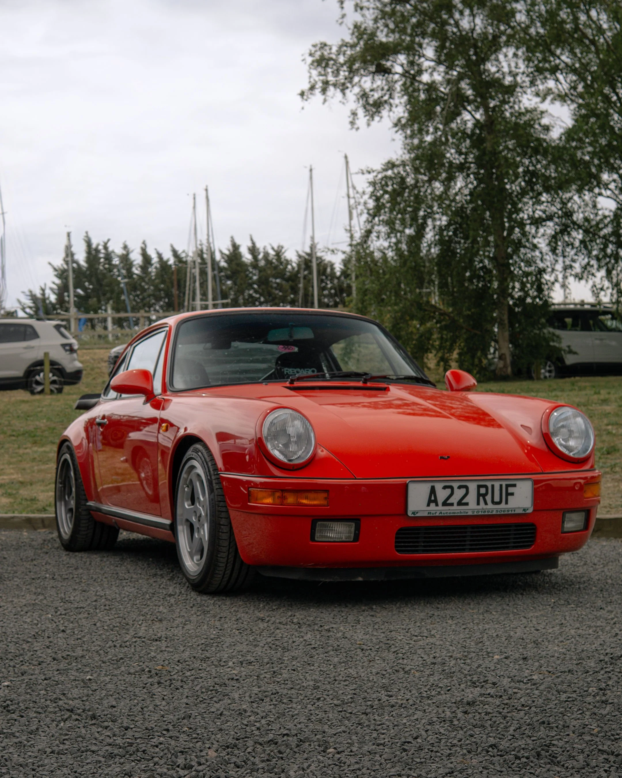 Red vintage sports car parked on gravel, with trees, sailboat masts, and other vehicles in the background under a cloudy sky.