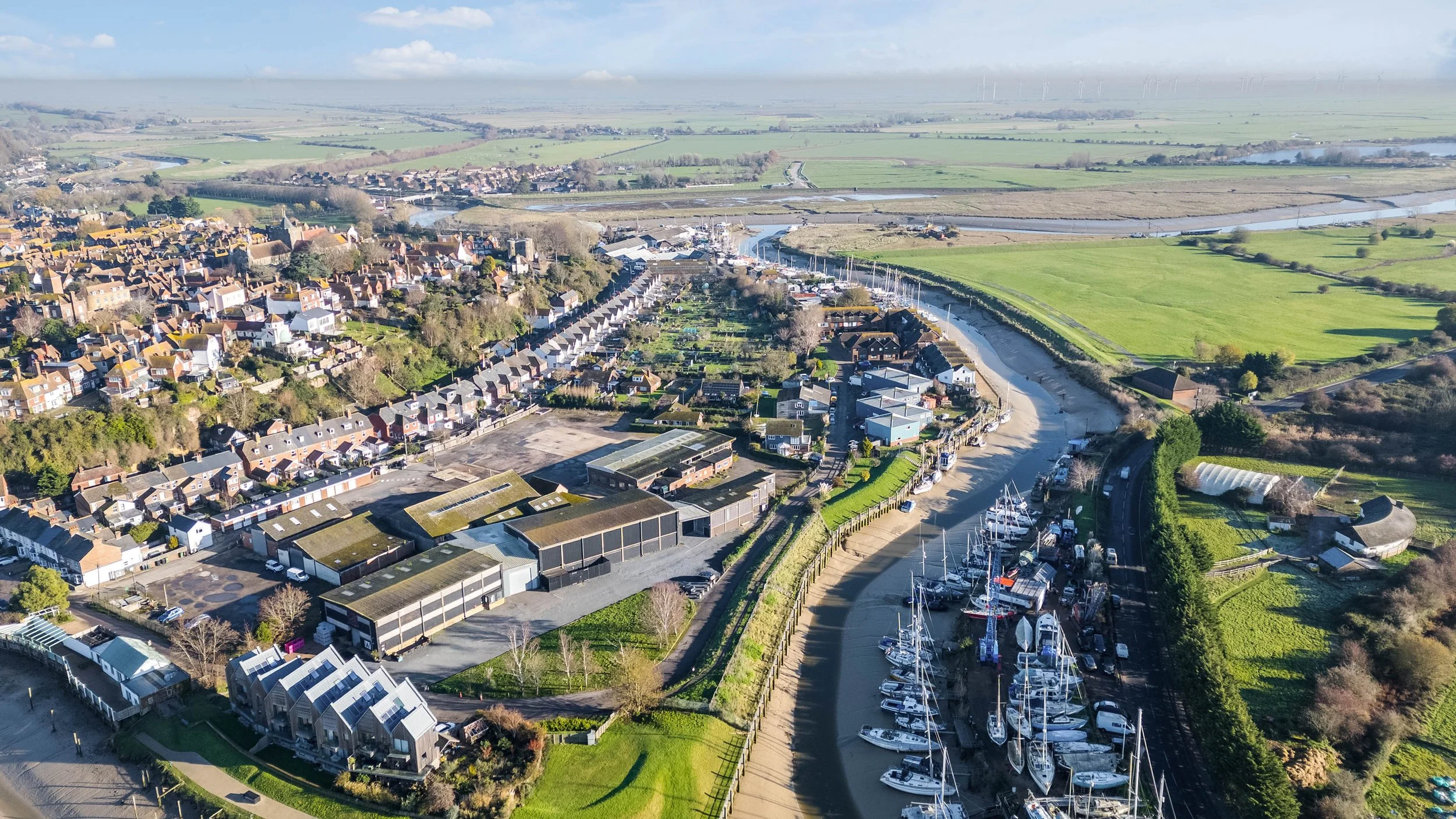 Aerial view of a coastal town with a marina filled with sailboats, residential houses, green fields, and a river in the background.
