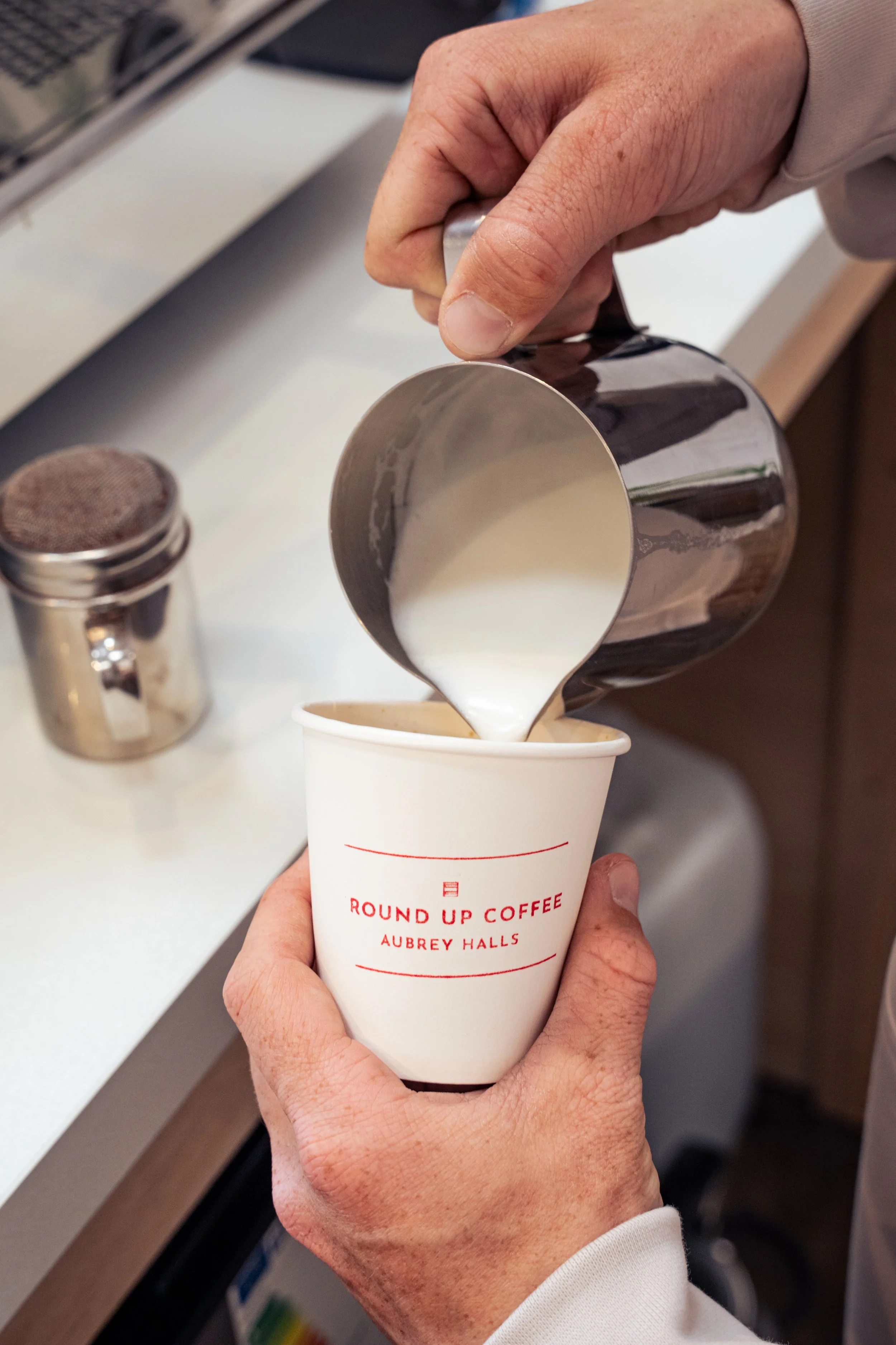 Person pouring milk into a paper coffee cup labeled 'Round Up Coffee Auburn Halls' in a coffee shop.