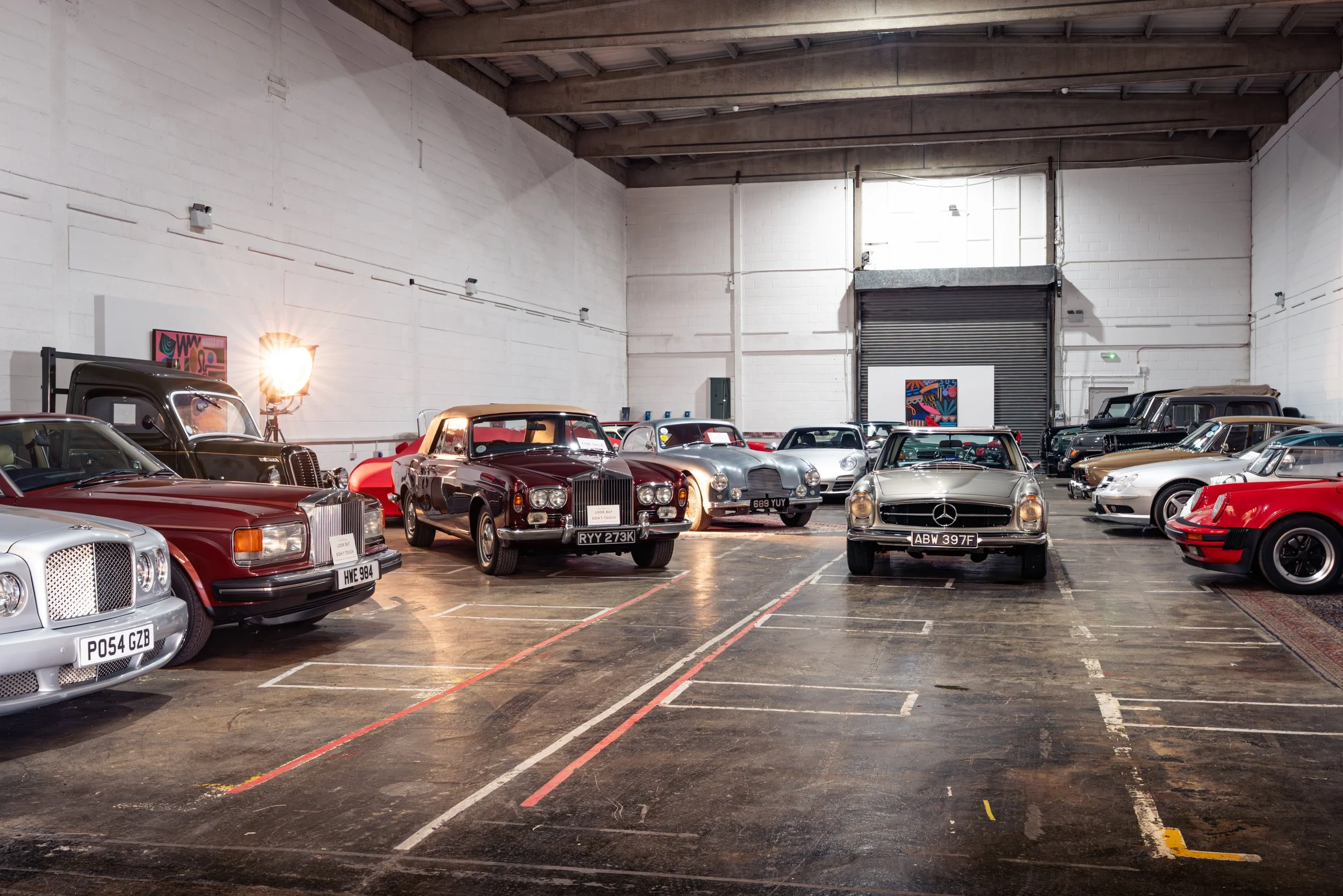Various vintage and modern cars parked inside a spacious warehouse car storage facility with a high ceiling, white brick walls, and artwork on the walls.
