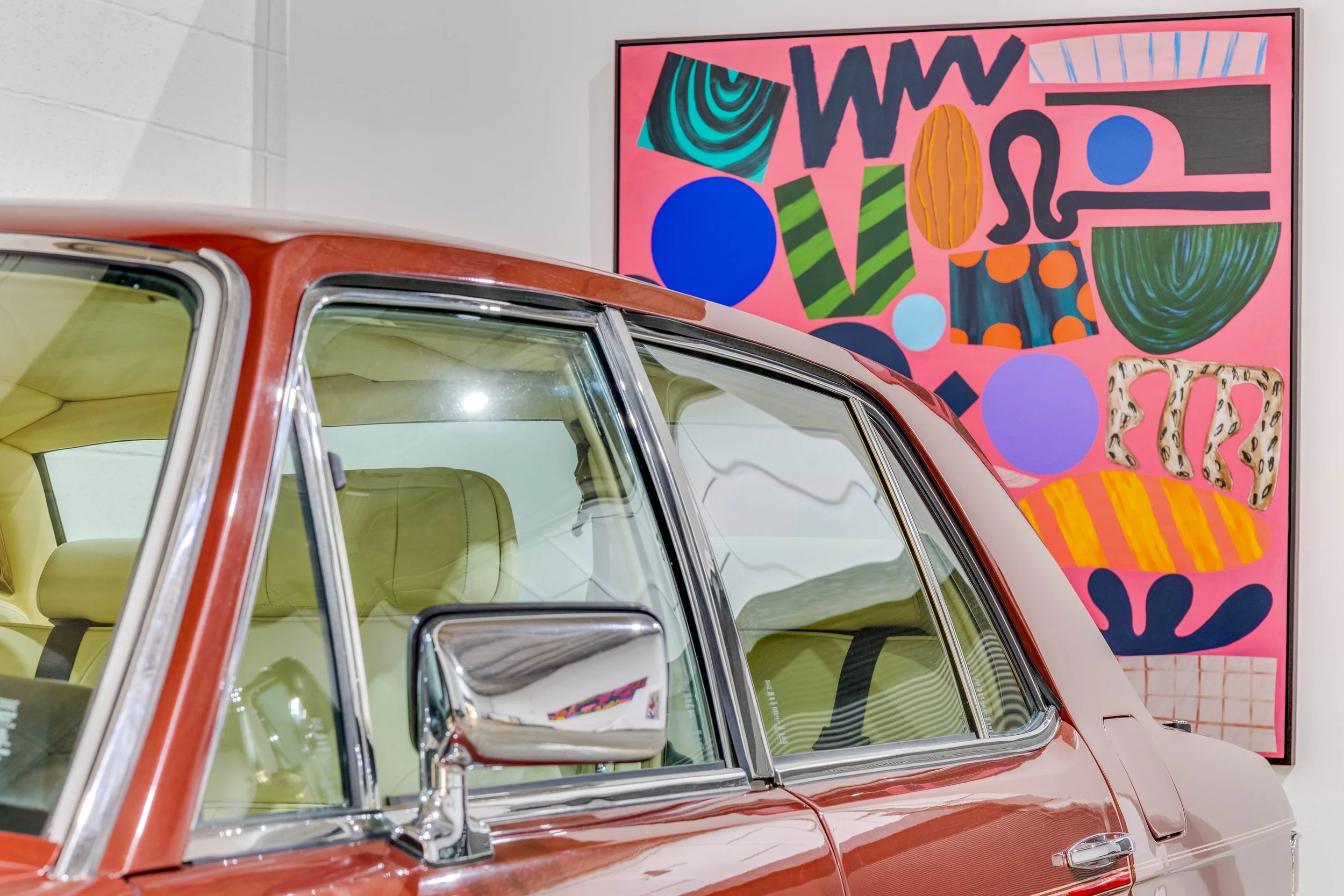 Close-up of a vintage car with a red and cream exterior, parked indoors, with an abstract colorful painting on the wall in the background.