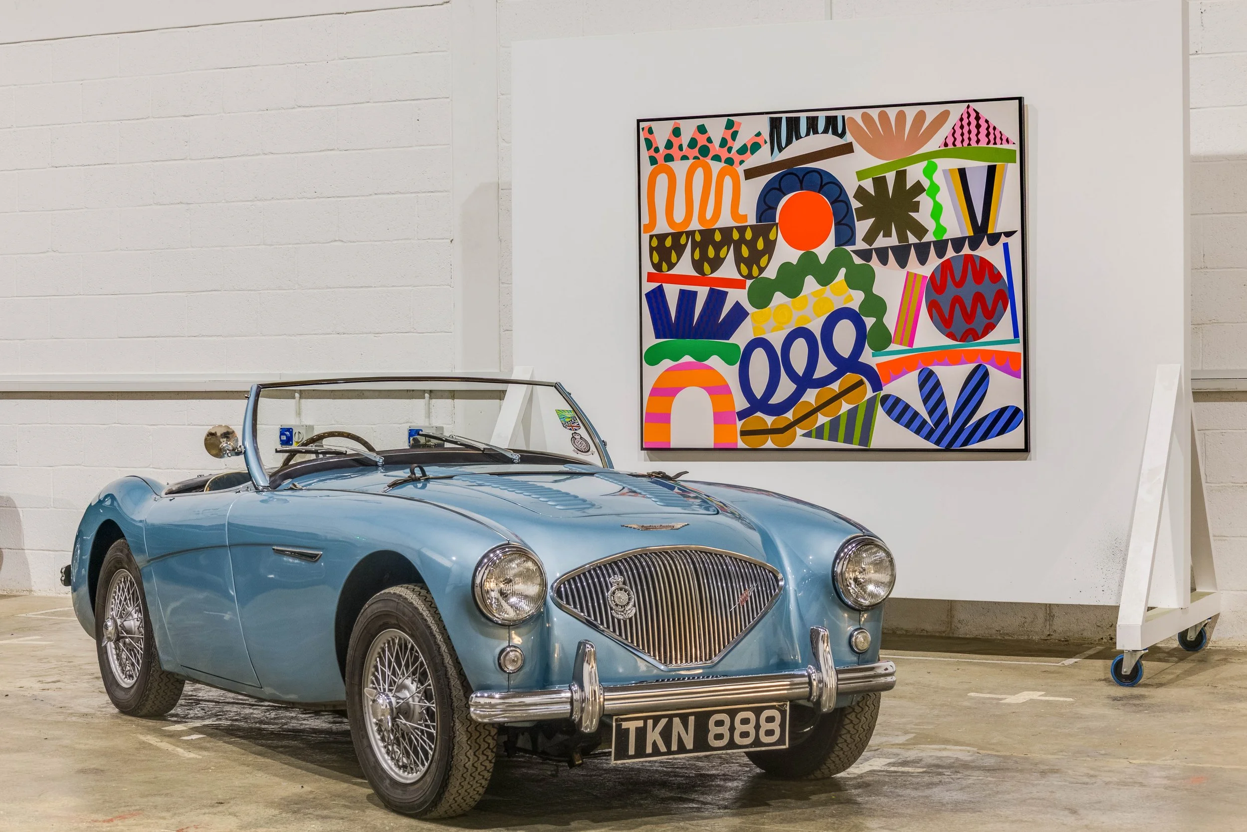 A vintage blue convertible car parked indoors next to a colorful abstract art piece on a white wall.