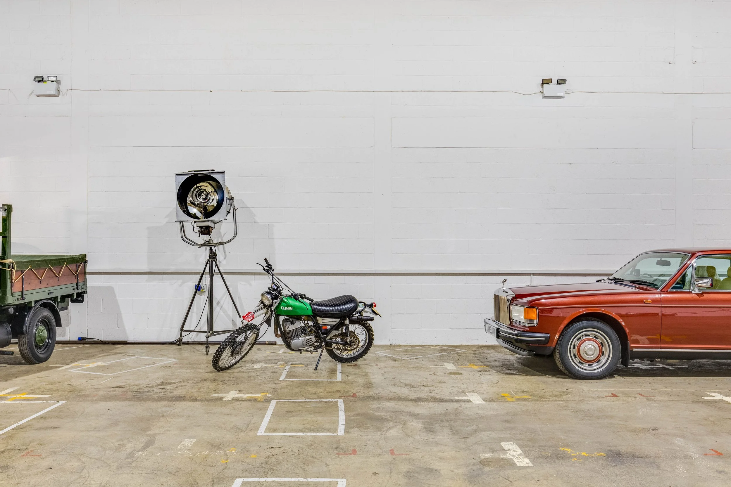 An indoor parking lot with a green motorcycle, an old red car, and part of a green truck, against a white wall with lighting fixtures.