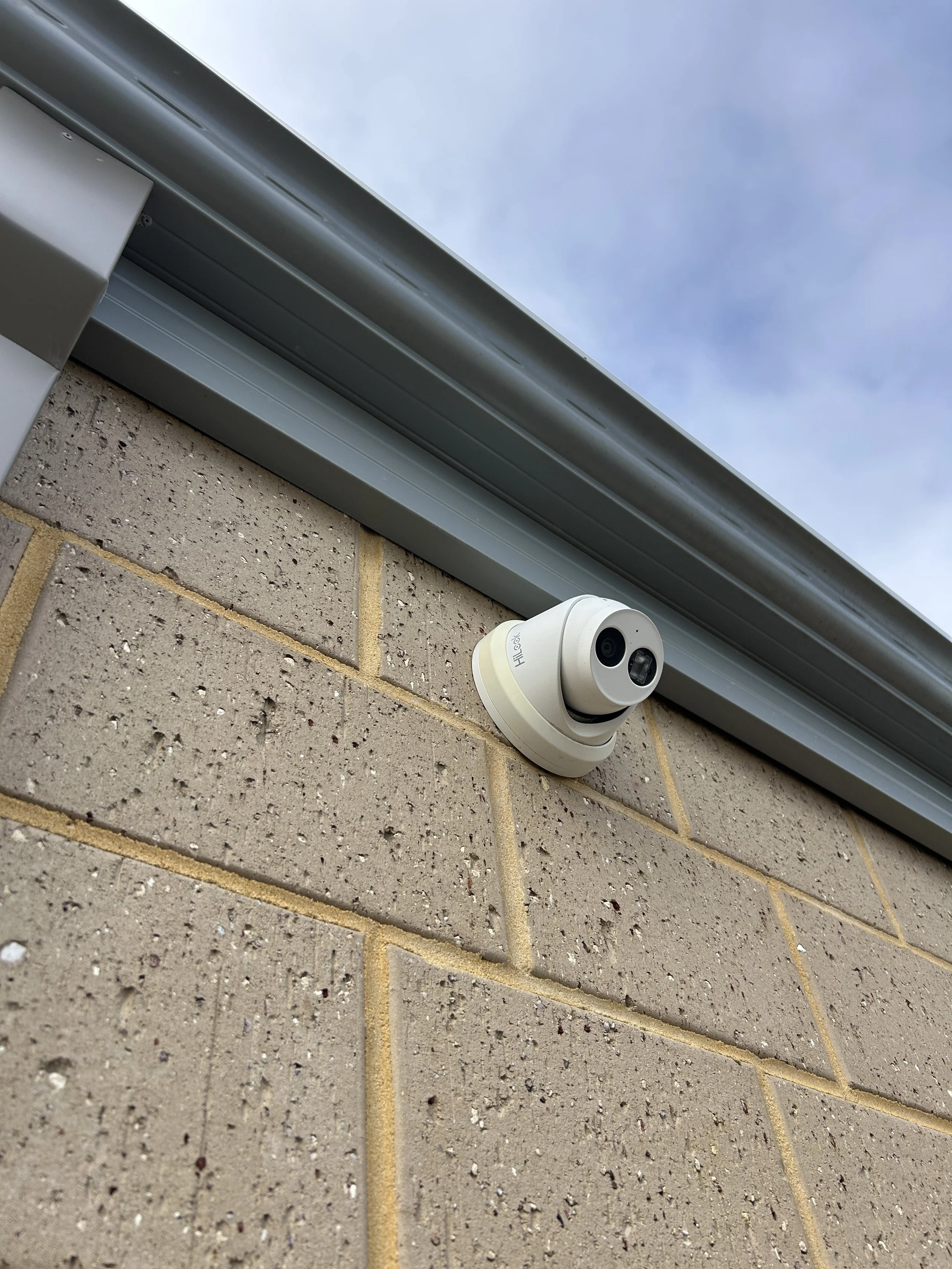 Close-up of a beige security camera mounted on a brick wall, with a metal guttering and a cloudy sky above.