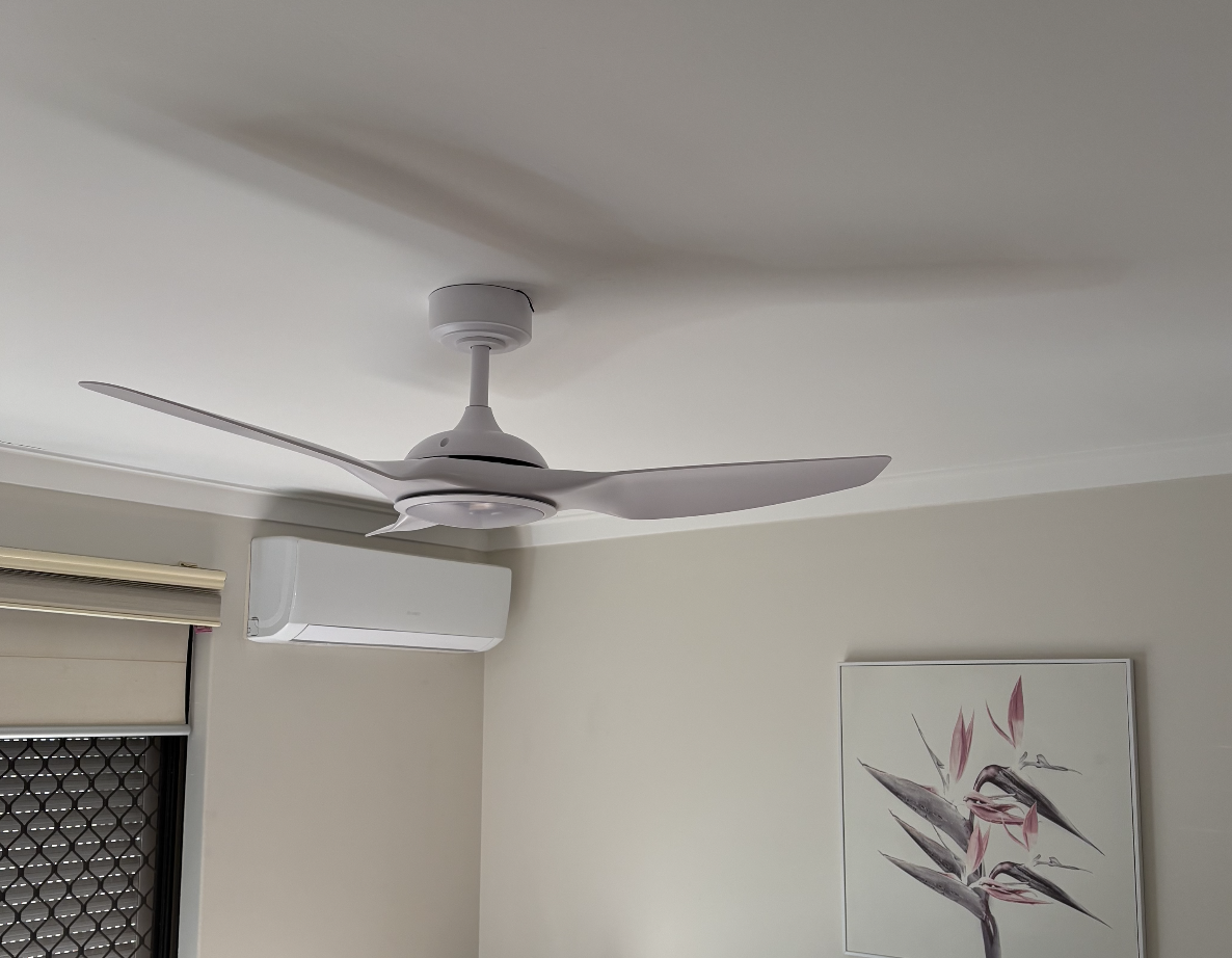 White ceiling fan with three blades in a room with light-colored walls. An air conditioning unit and a framed floral artwork are visible on the wall.