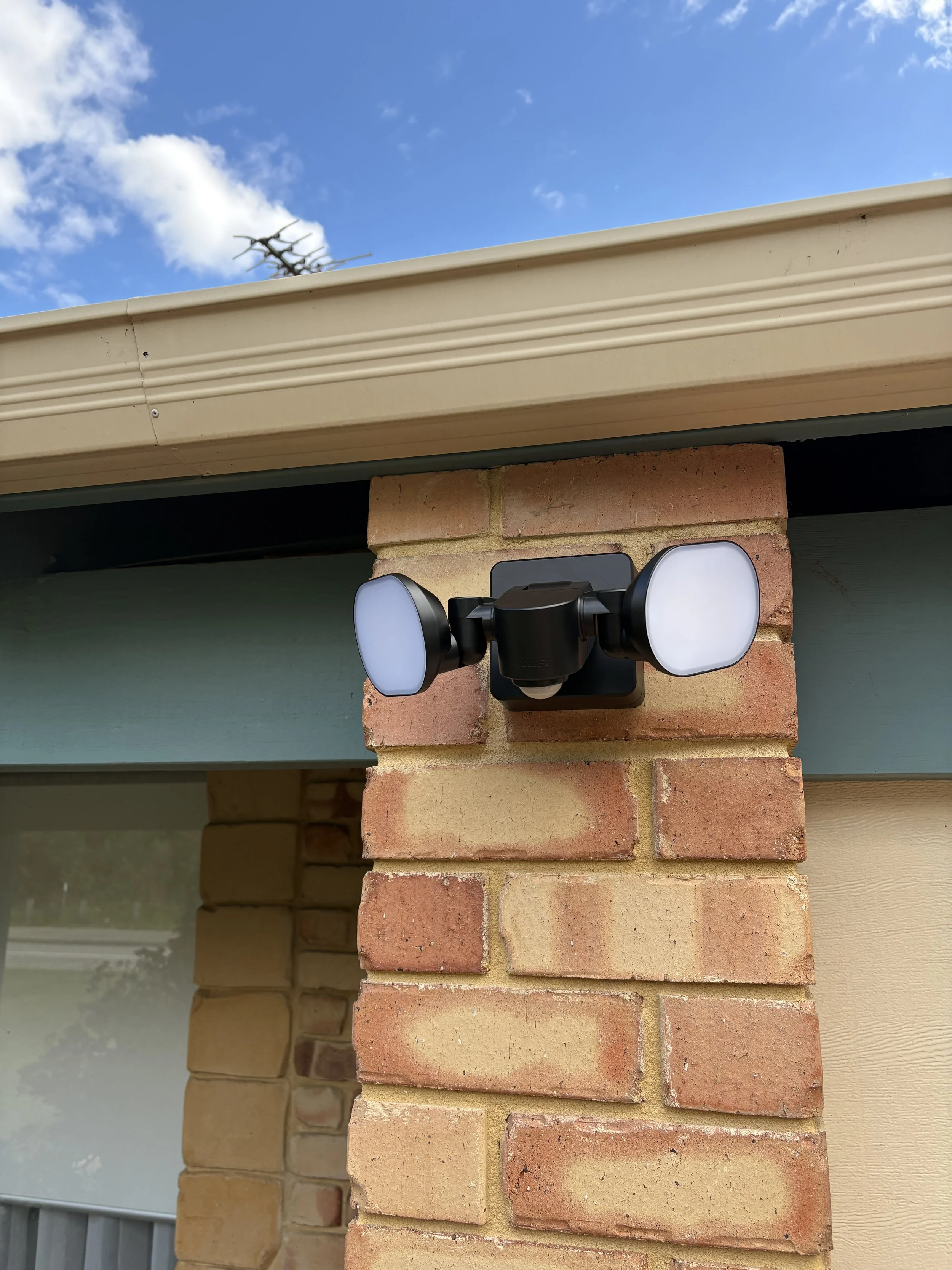 Close-up of a brick column with a mounted outdoor camera, part of a house exterior under a blue sky with some clouds.