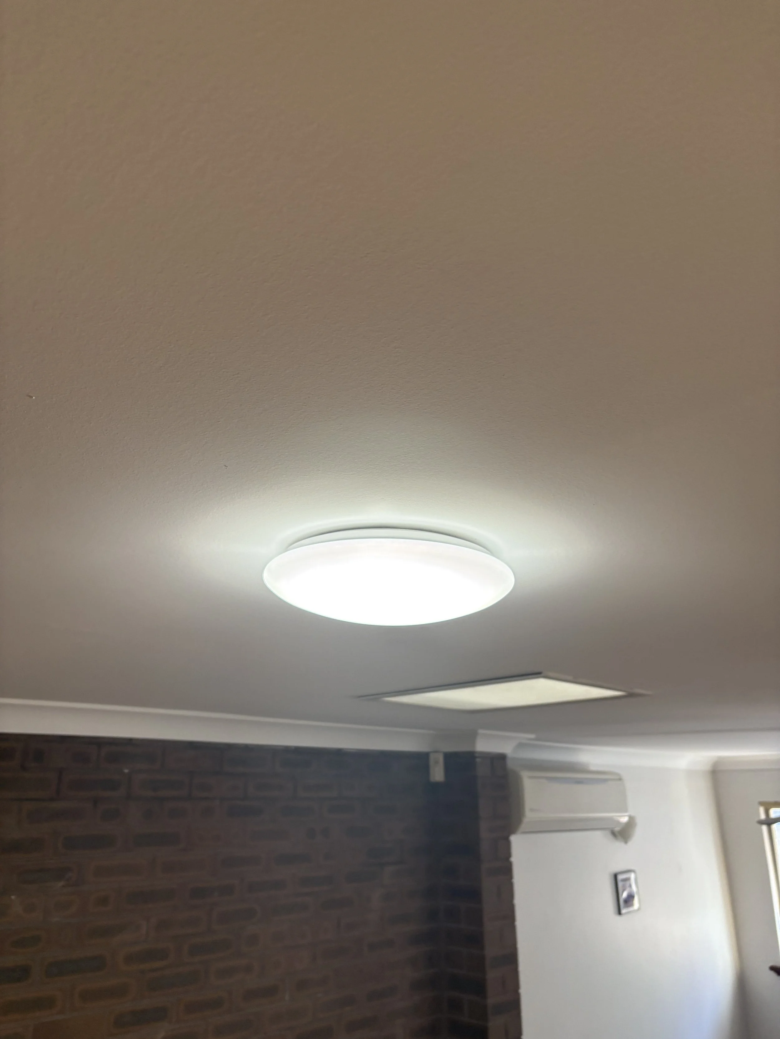 Ceiling with two round light fixtures, one bright and white, and a white air vent, with part of a brick wall and white wall with framed picture and white air conditioning unit visible.