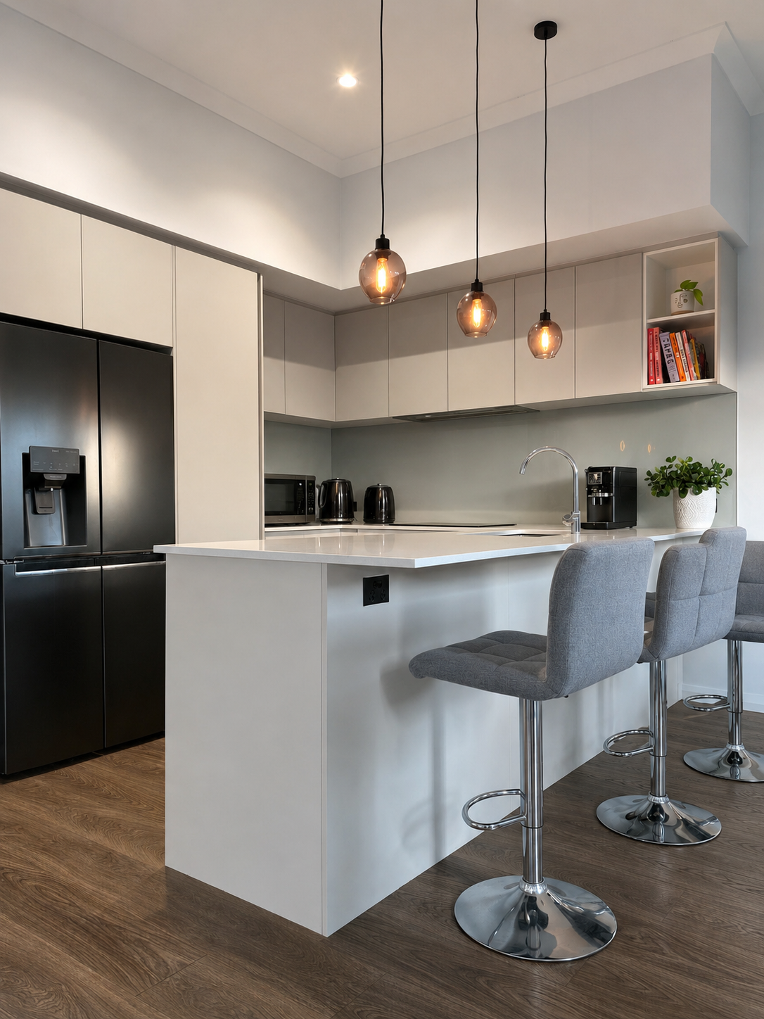 Modern kitchen with black refrigerator, white cabinets, kitchen island with grey bar stools, pendant lighting, coffee maker, microwave, black kettles, potted plant, and books on open shelves.