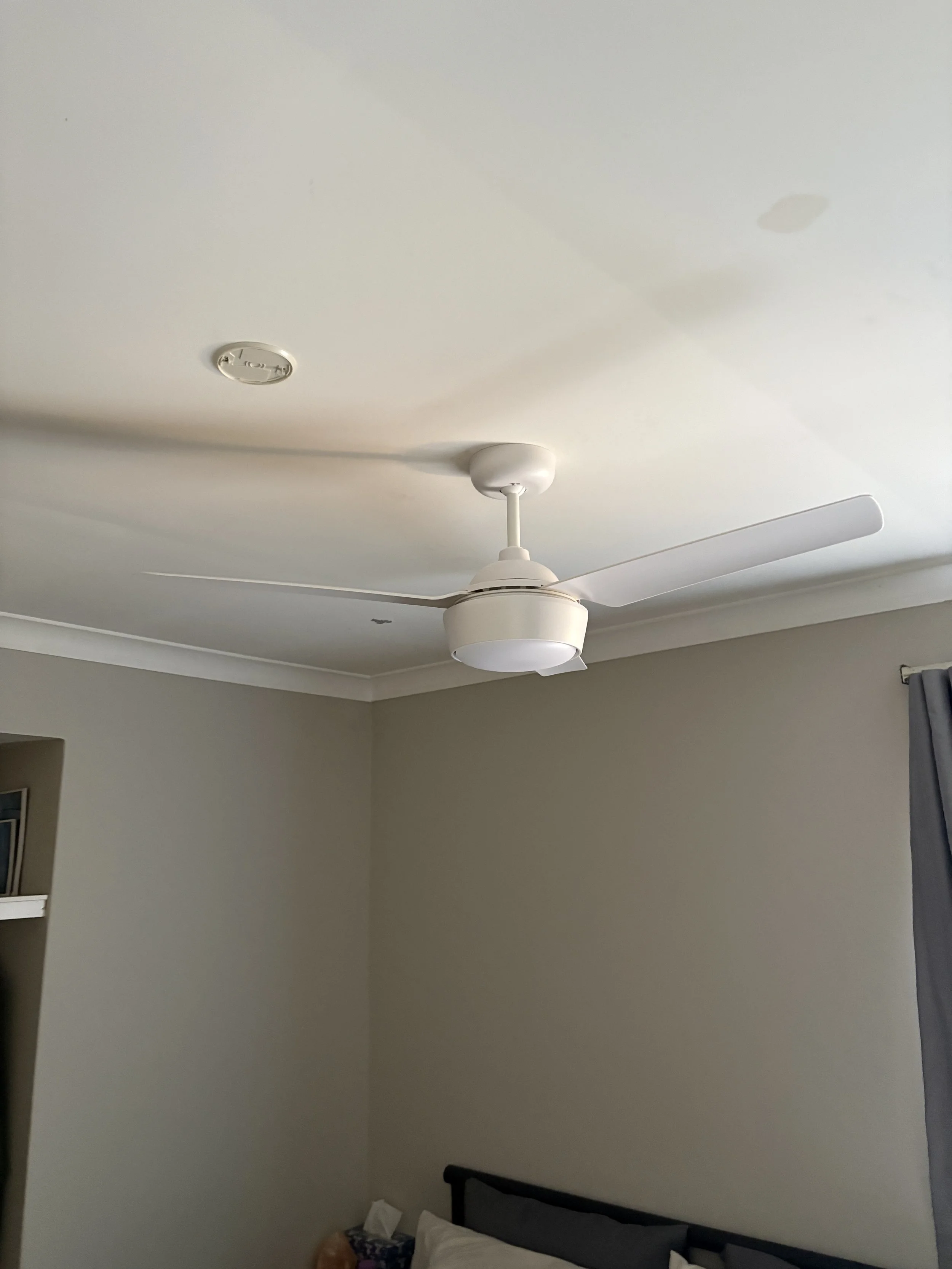 A bedroom with beige walls, a white ceiling fan, and a ceiling light fixture. Part of a bed is visible at the bottom of the image, along with a trash bin and some objects on the floor.