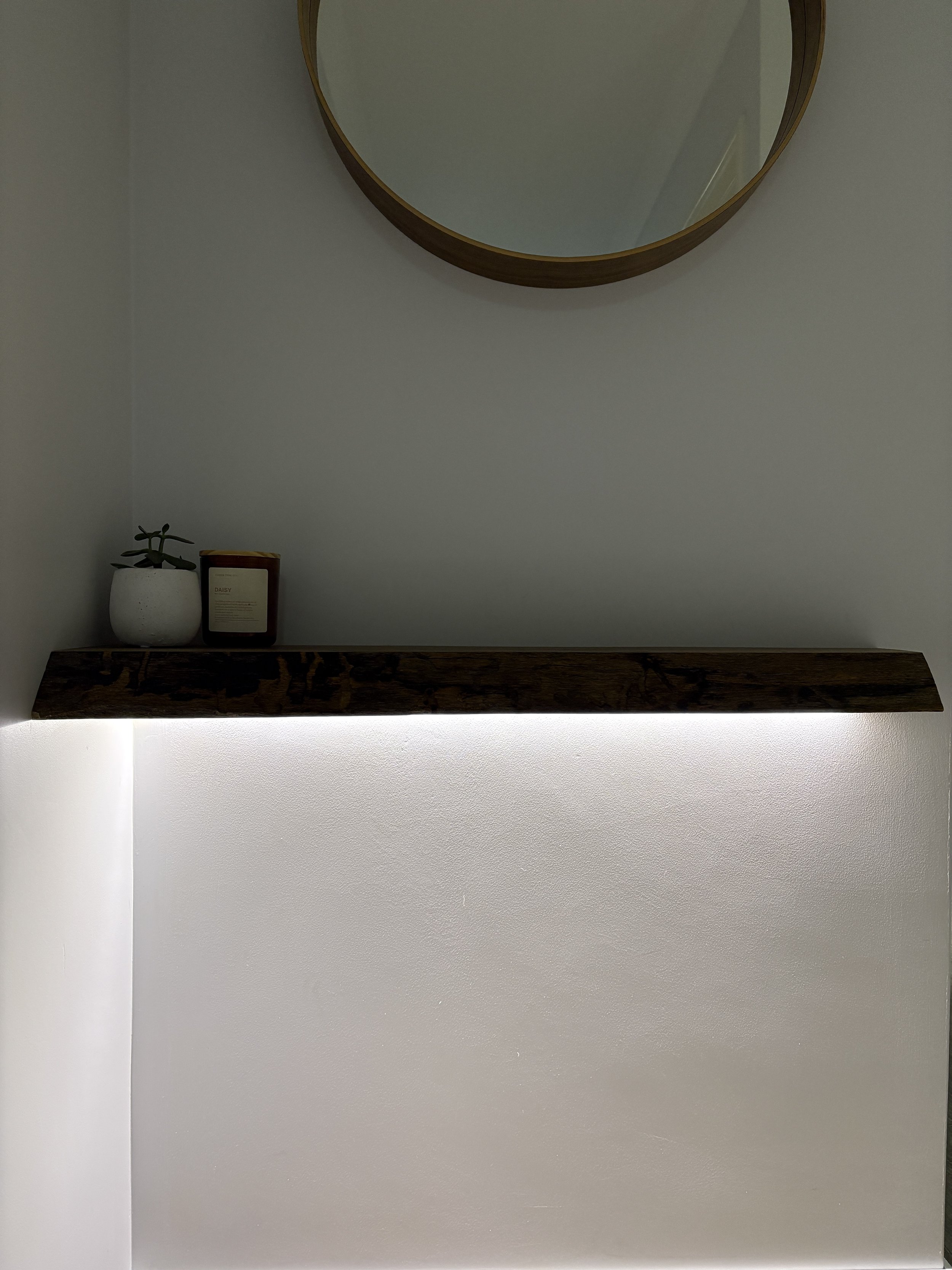 A white potted plant and a candle in a box on a dark wooden shelf against a wall, with a large round mirror above.