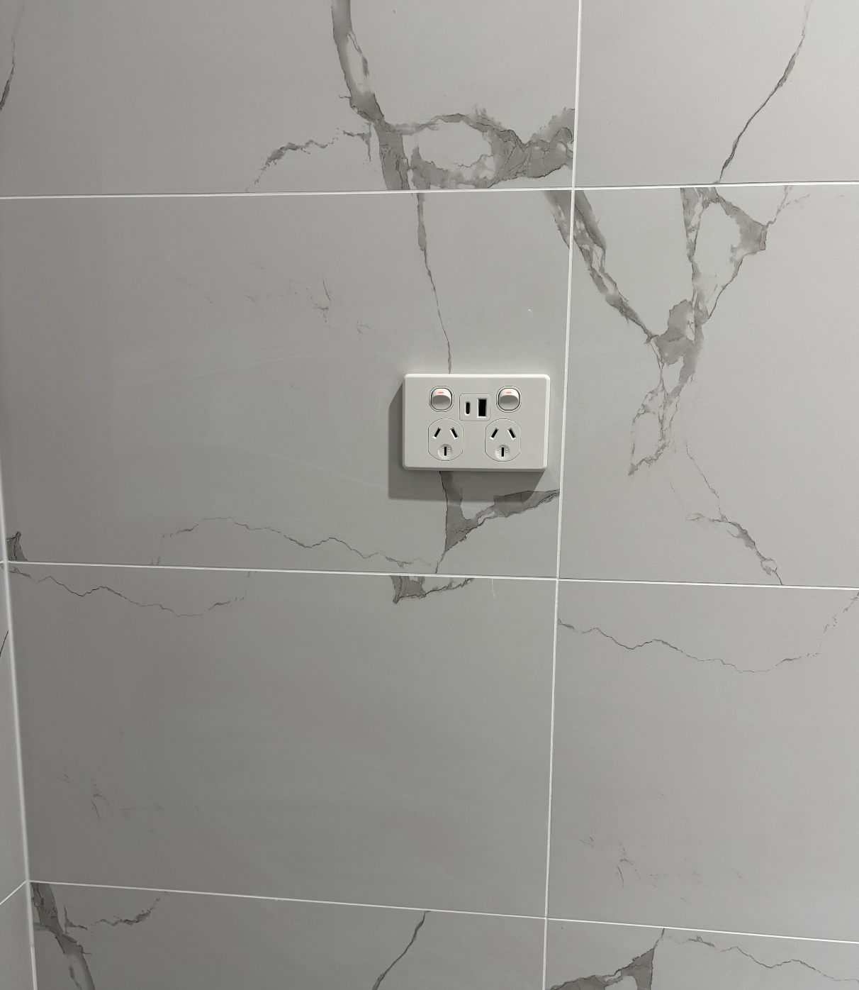 A white wall socket with multiple plugs and switches attached to a marble-patterned tile wall.