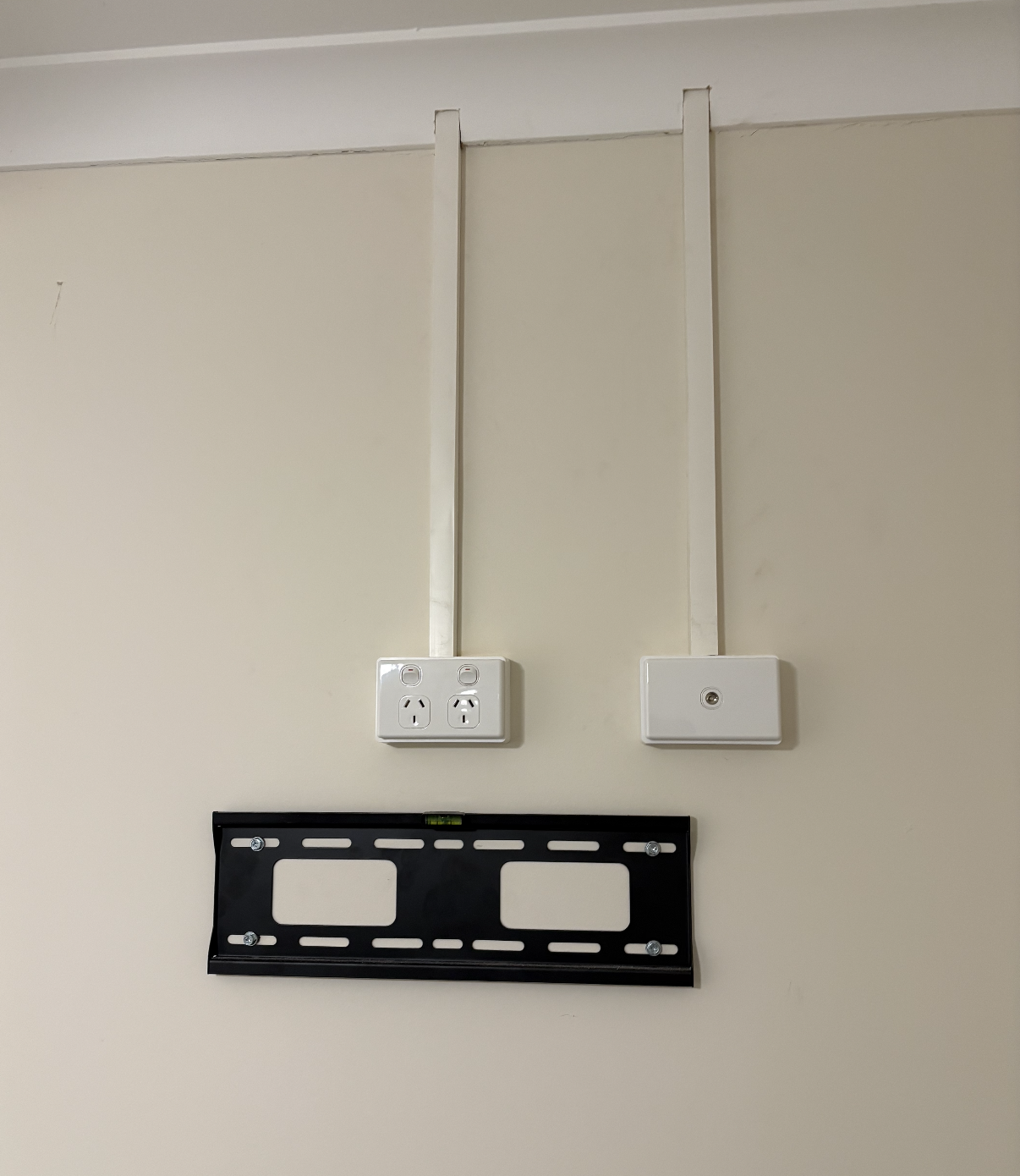 Wall with two electrical outlets, one with power switches, and a black mounting bracket for a TV or monitor on a beige wall.