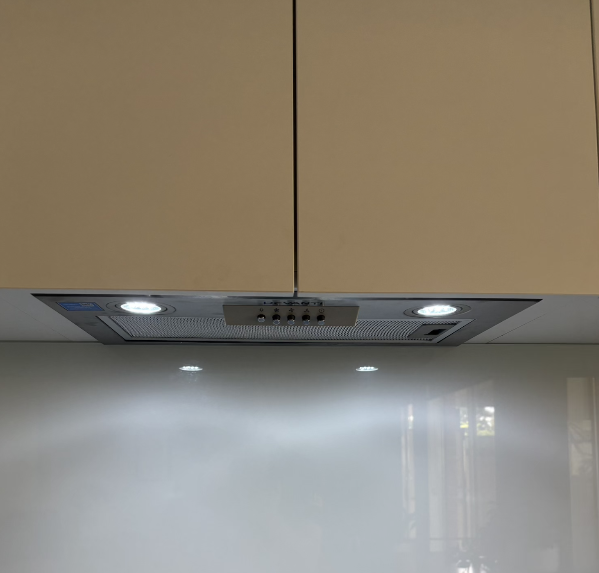 Close-up of a beige kitchen range hood with built-in lights and control buttons, mounted on a wall with brown cabinets above.
