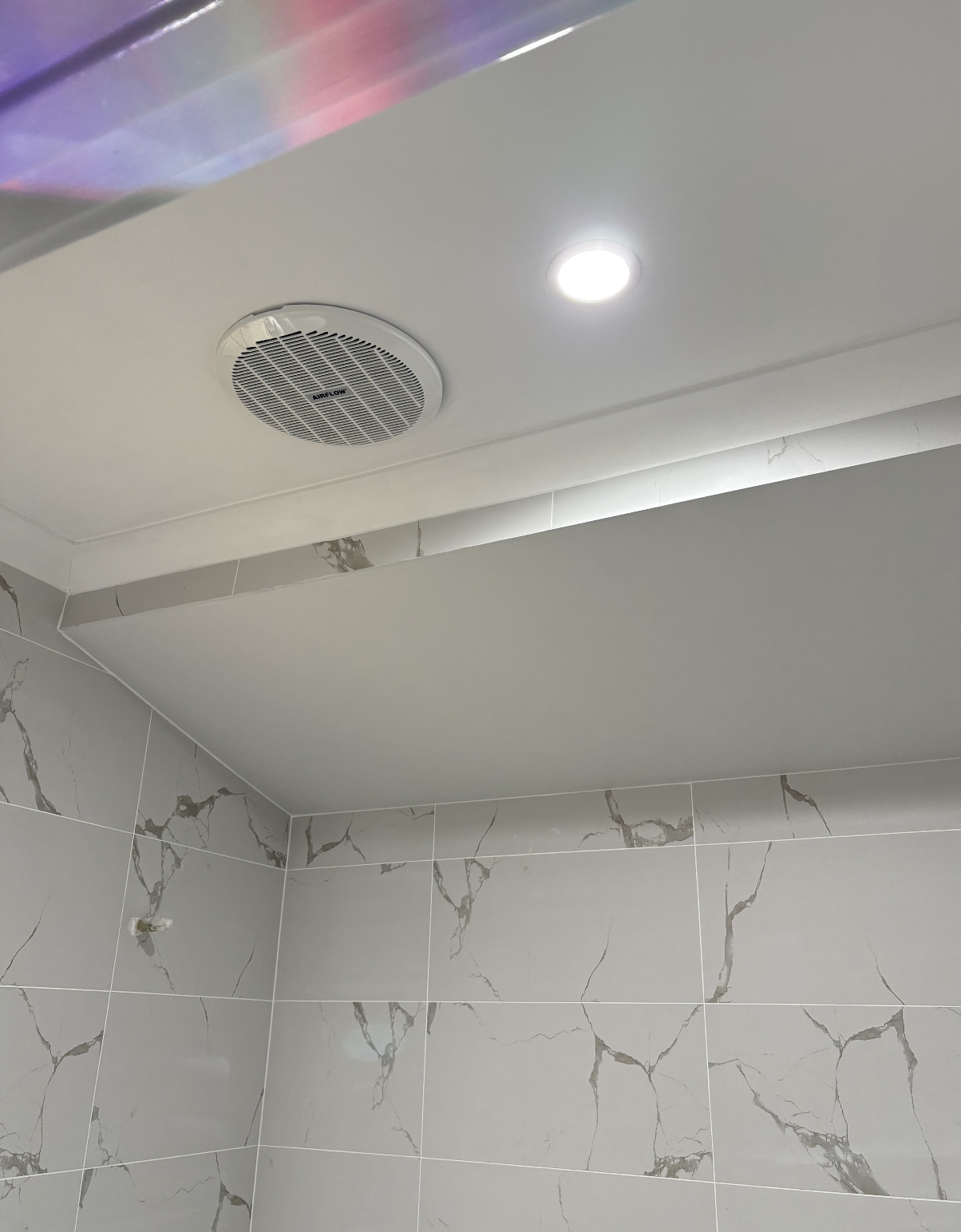 Ceiling with a round ventilation fan and a recessed light in a bathroom with marble-patterned gray tiles.