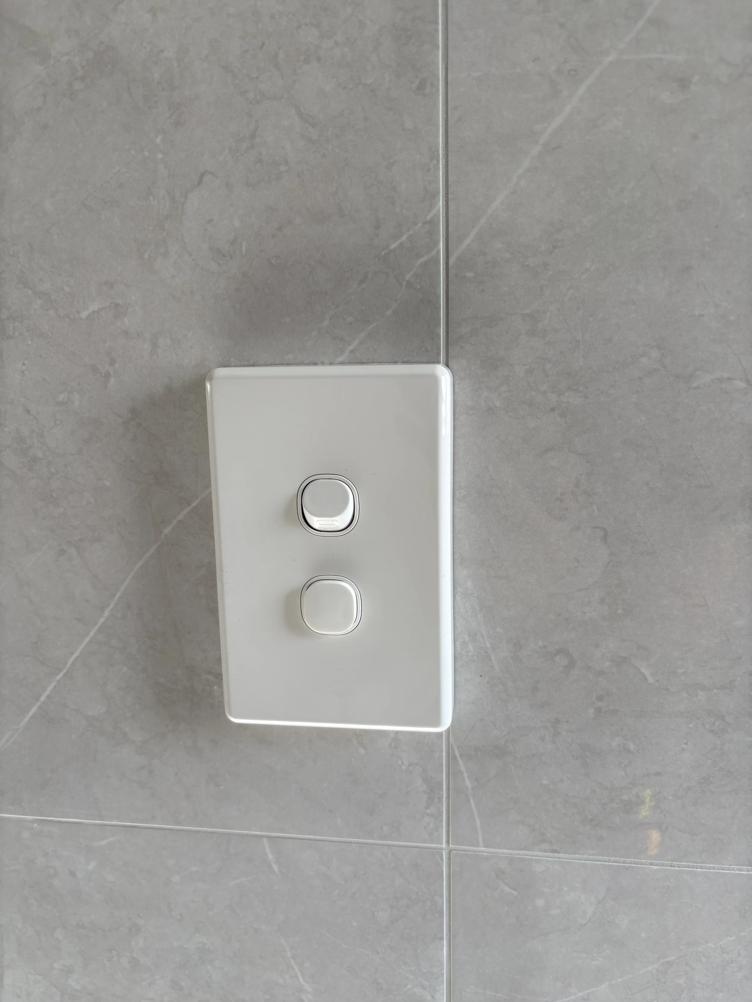 Close-up of a white light switch with two buttons mounted on a gray tiled wall.