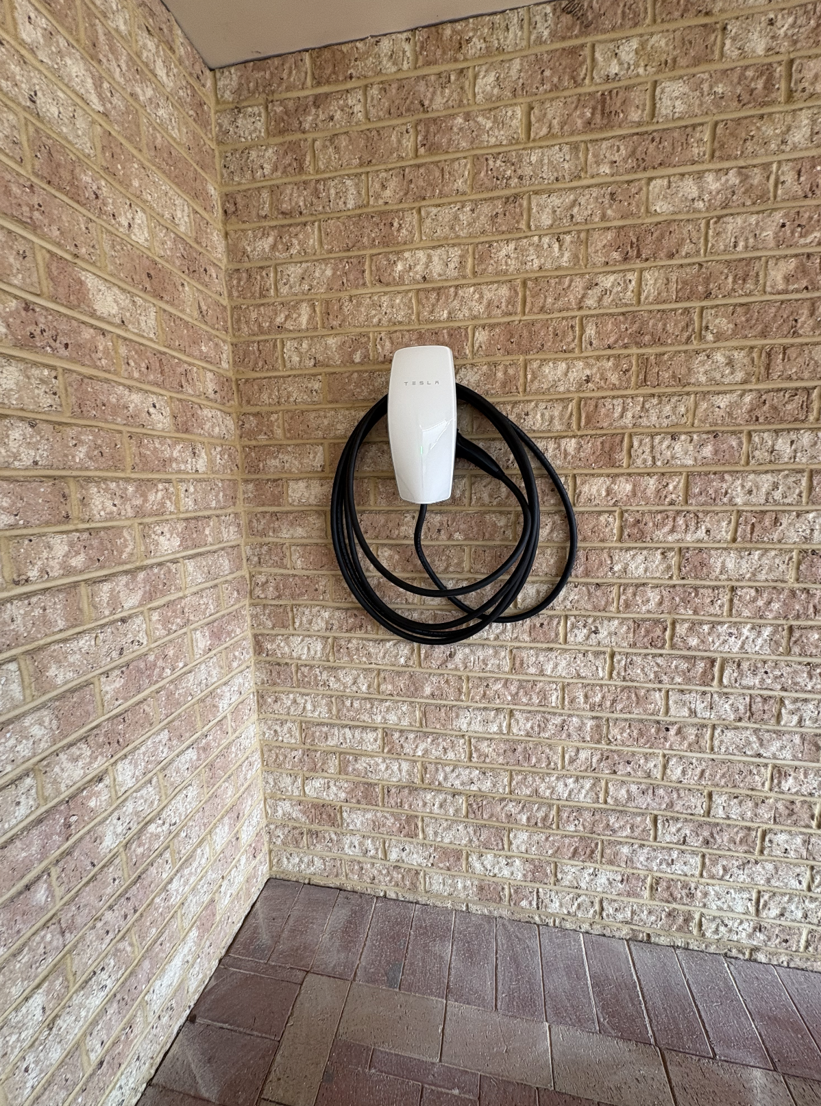Electric vehicle charging station mounted on a brick wall in an outdoor corner, with a plugged-in charging cable hanging below.