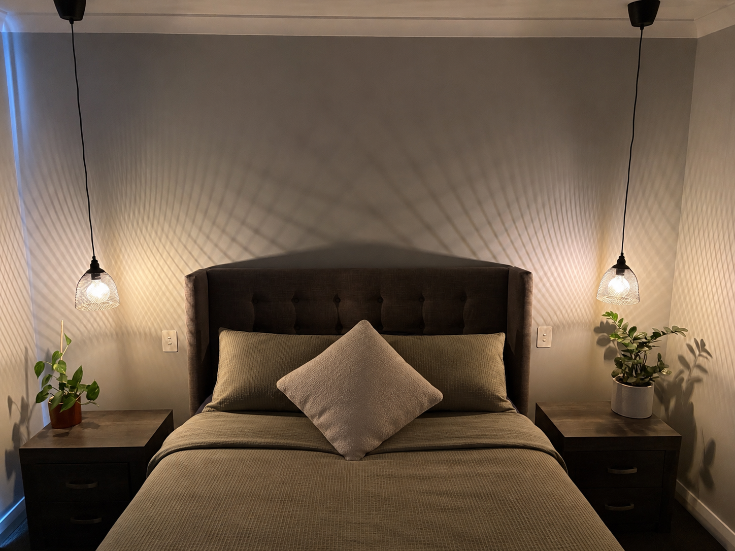 A bedroom with a bed, two nightstands, pendant lights, and potted plants, with shadows creating a geometric pattern on the wall behind the bed.