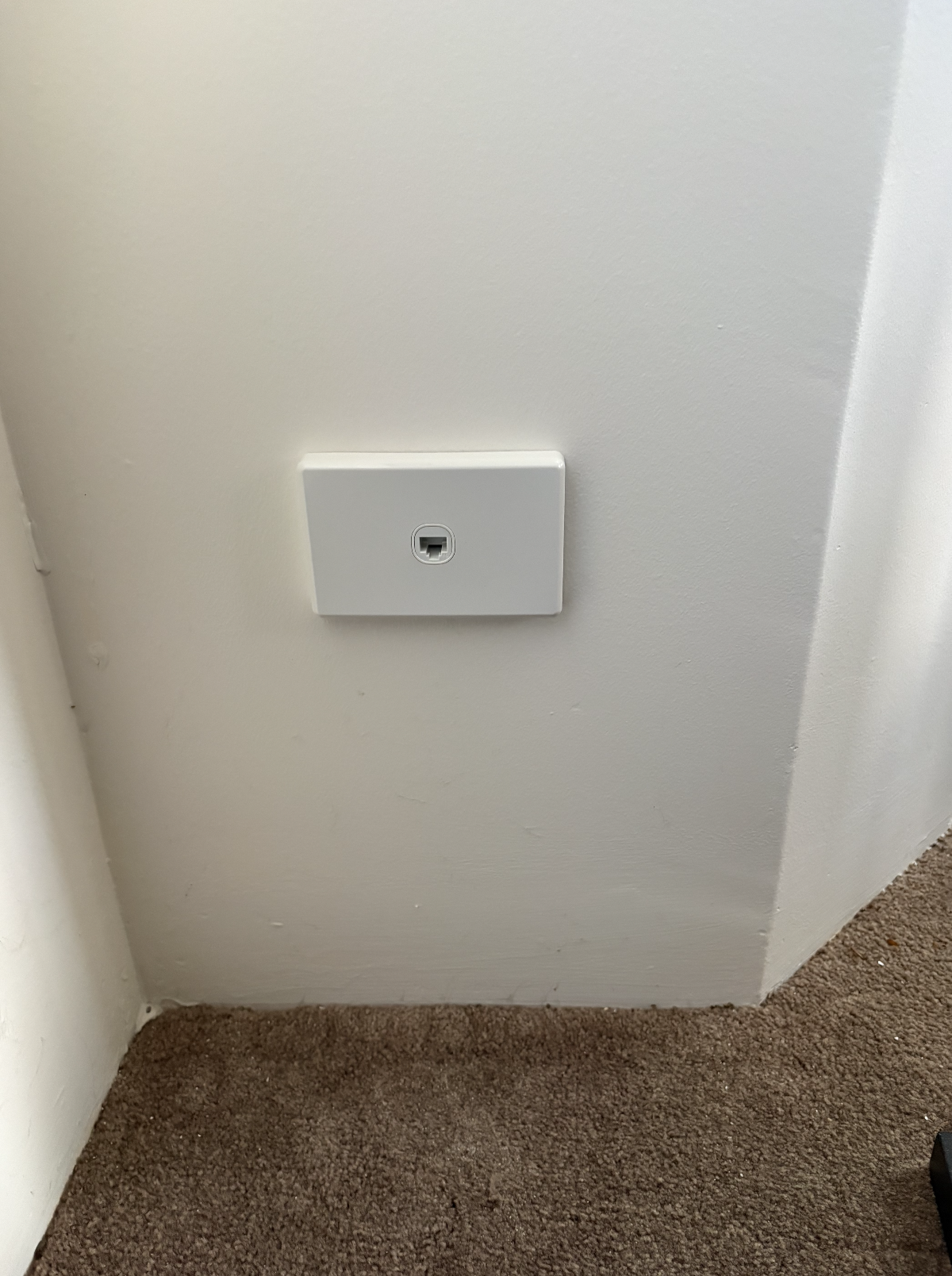 Close-up of a white wall vent or outlet cover on a cream-colored wall near brown carpeted flooring.