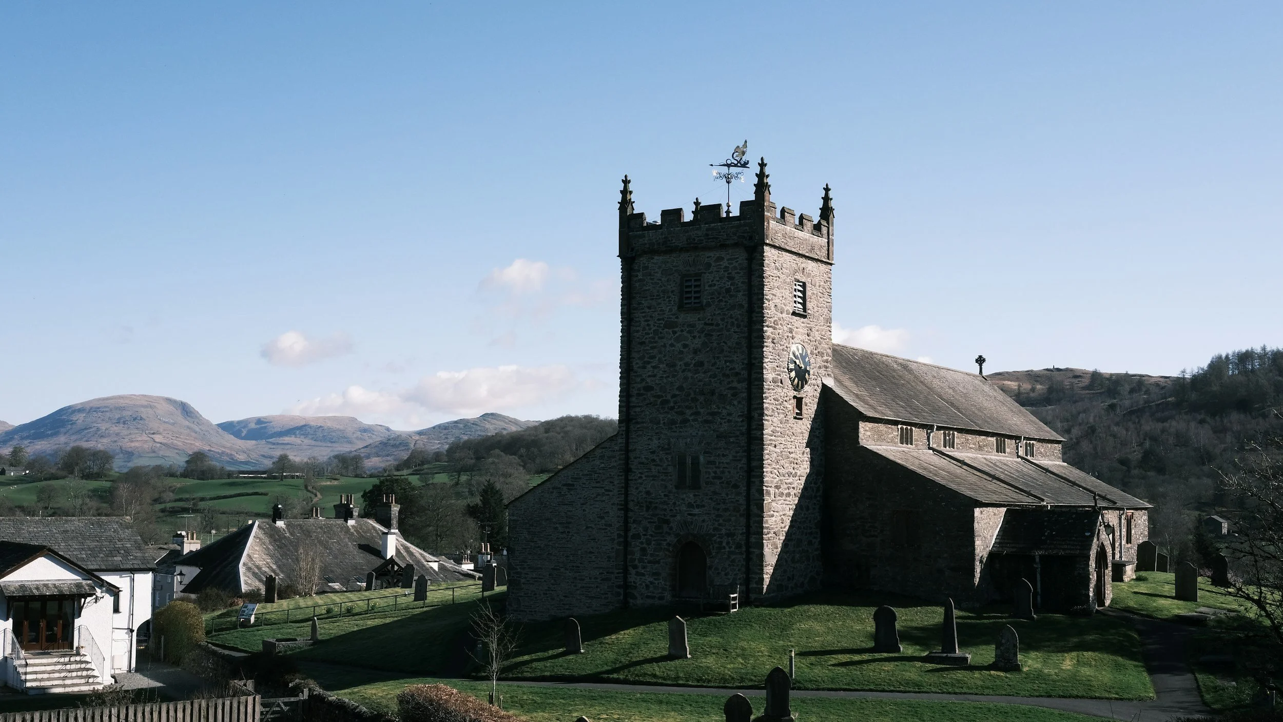 Historic stone church with a tall square clock tower under a blue sky, surrounded by a small hillside churchyard.