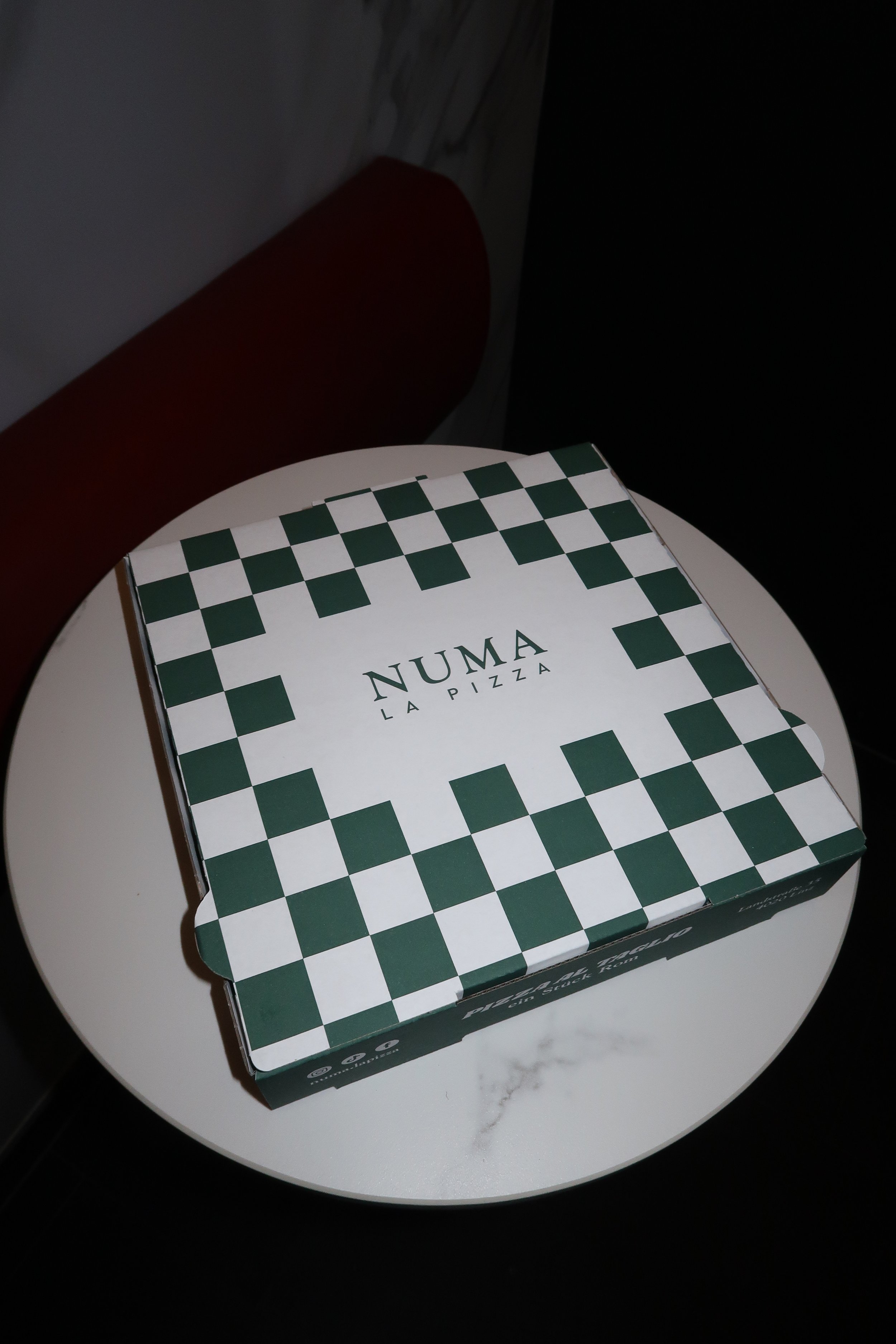 Pizza-Box mit grün-weißen Karomuster und 'NUMA LA PIZZA'-Schrift auf einer weißen runden Tischplatte.