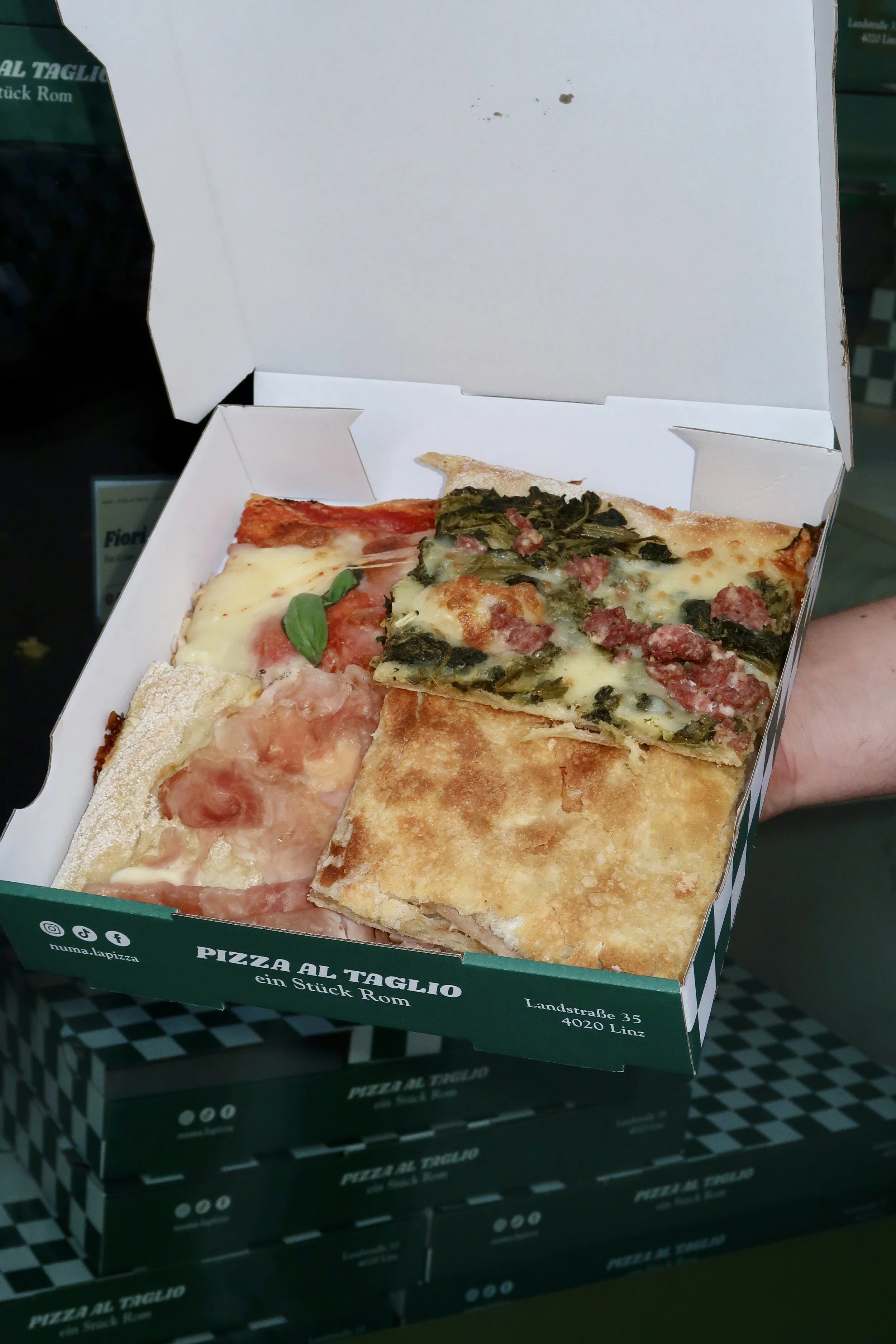 Offene Pizzabox mit vier verschiedenen Pizzastücken, darunter eine mit Tomaten, eine mit Spinat und Speck, eine mit Schinken und eine mit Käse, auf einem schwarzen Tisch.